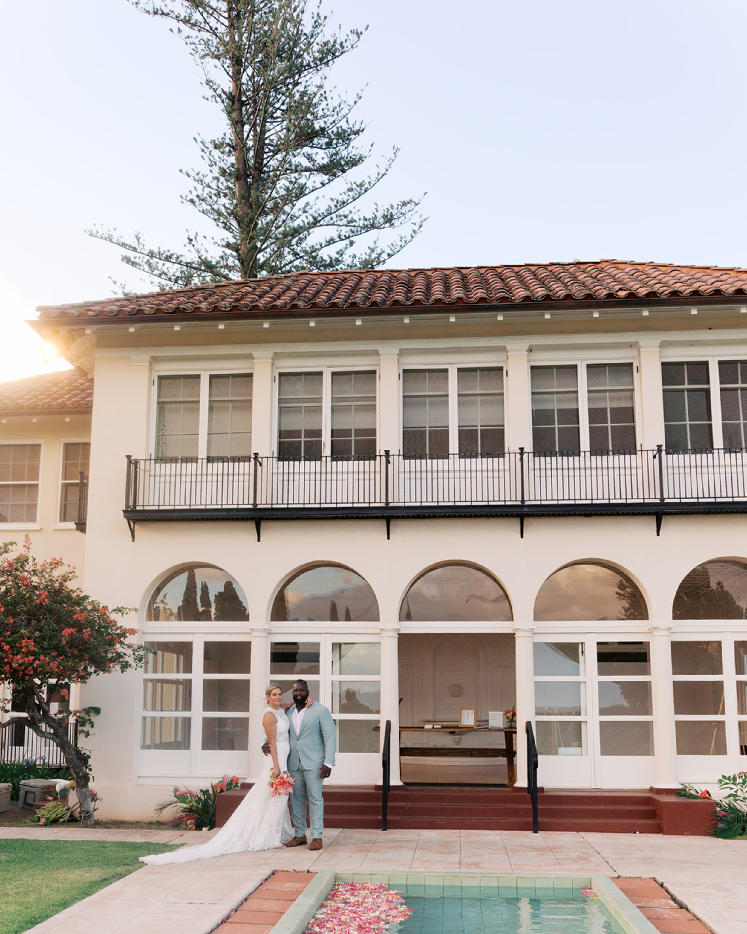 A couple standing outside a white building with a red tiled roof, large windows, and a balcony, decorated for a wedding. They are smiling and standing on steps leading to the entrance, with a pool filled with flower petals in the foreground. The coup