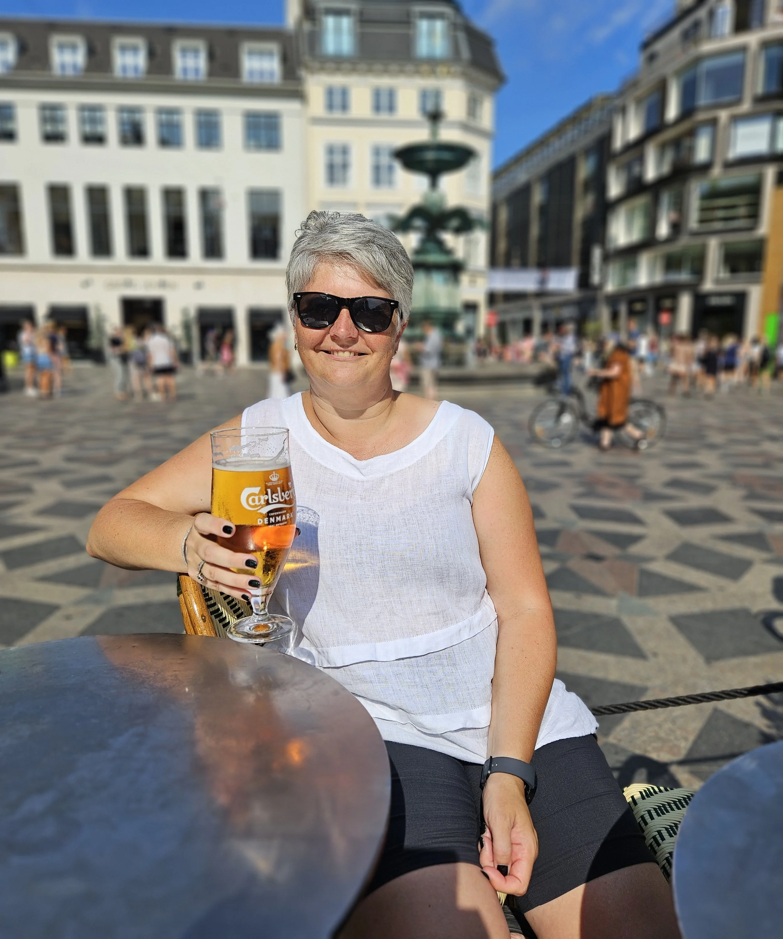 A woman with short gray hair, wearing sunglasses and a sleeveless white top, smiling and holding a glass of beer at an outdoor cafe in a European city square with historic buildings and a fountain in the background on a sunny day.