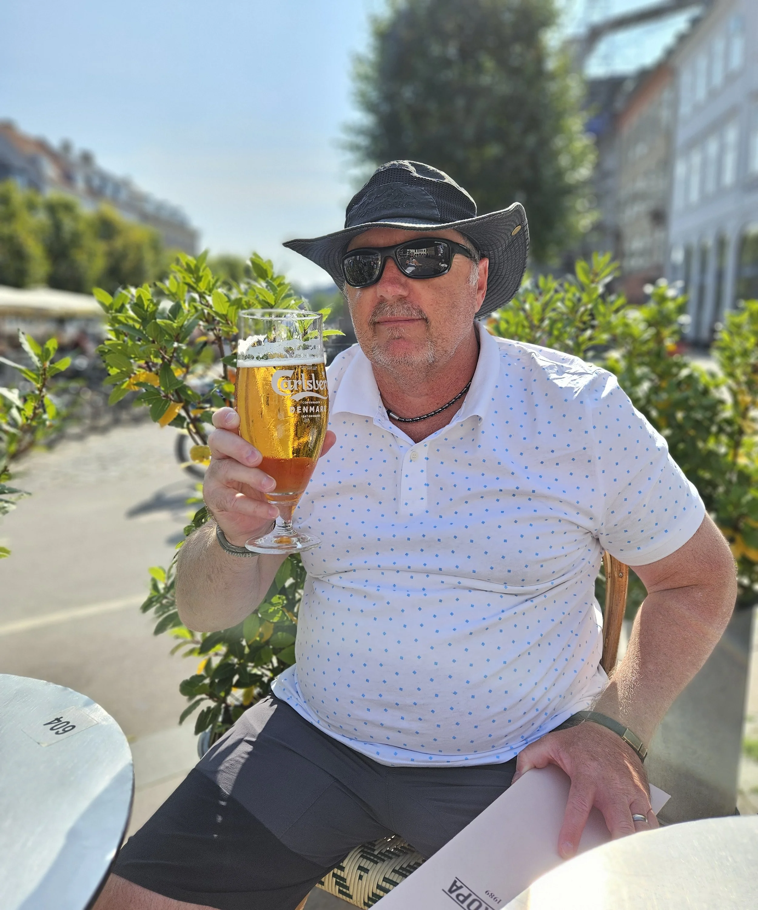 A man sitting outdoors holding a glass of beer, wearing a wide-brimmed hat and sunglasses, with a background of green bushes, buildings, and a clear blue sky.
