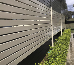 A close-up side view of a beige horizontal aluminium slat fence with a row of green hedges at its base, under a cloudy sky.