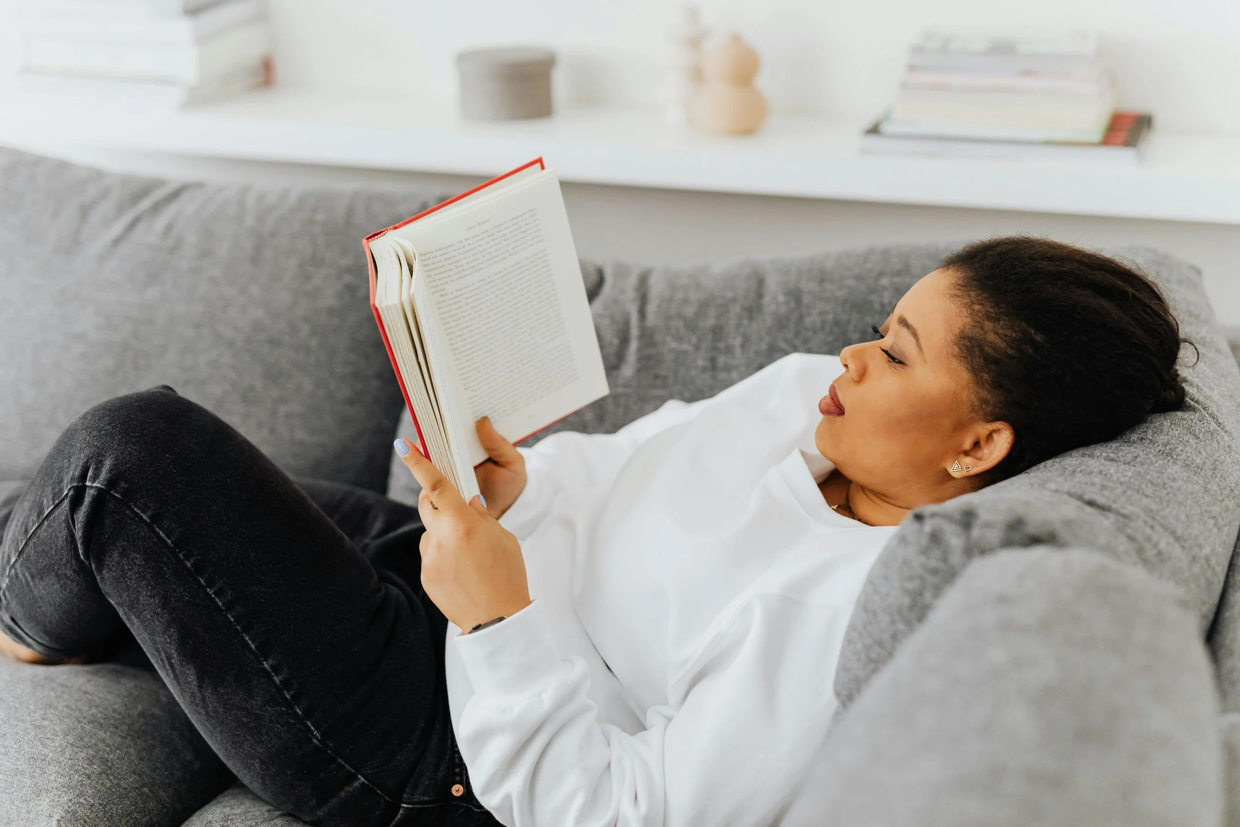 A woman with dark hair lying on a gray sofa, reading a book in a bright living room.