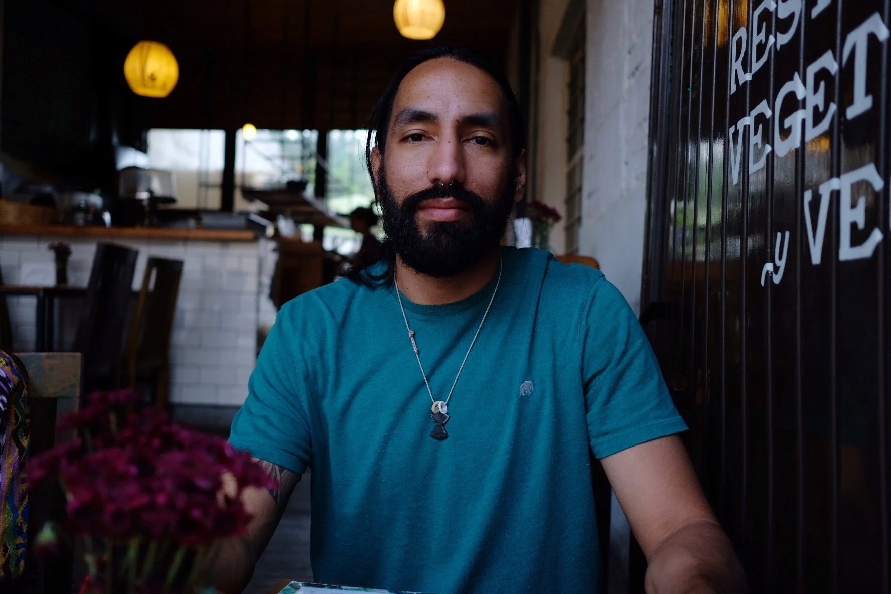 A man with long black hair, a beard, wearing a teal T-shirt and a necklace, sitting at a table in a restaurant with warm lighting and decor, including flowers and a sign with the words 'REAS, VE' visible.