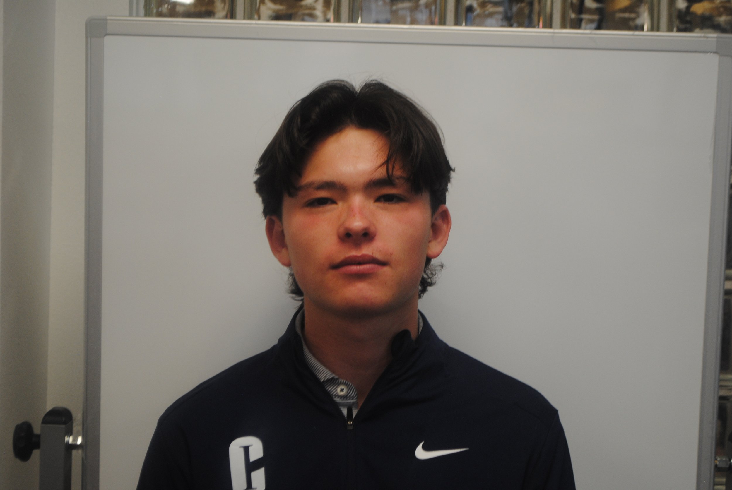A young man with dark hair and a neutral expression, wearing a black Nike jacket with a white logo, standing in front of a whiteboard.
