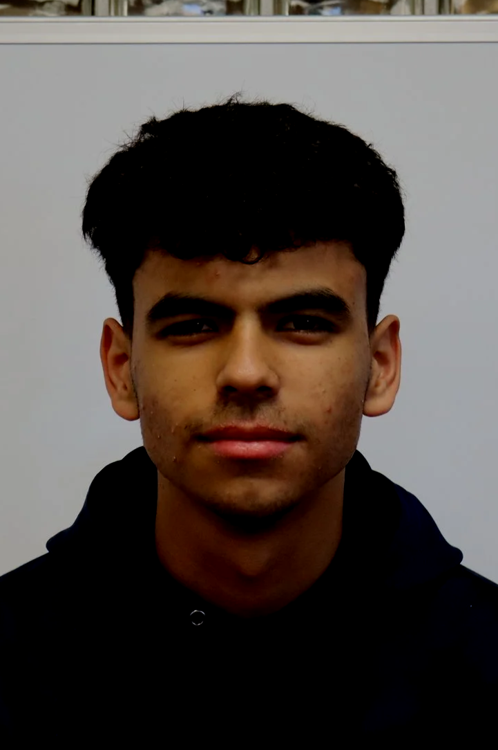 Portrait of a young man with short dark hair, wearing a black hoodie, standing against a plain light-colored background.