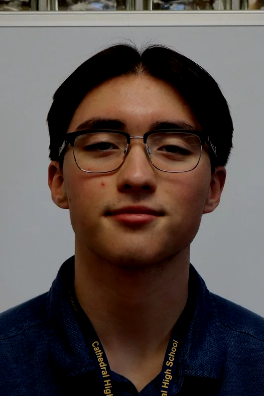 A young man with black hair, glasses, and a slight smile wearing a navy blue shirt and a lanyard that says "Cathedral High School" against a plain background.