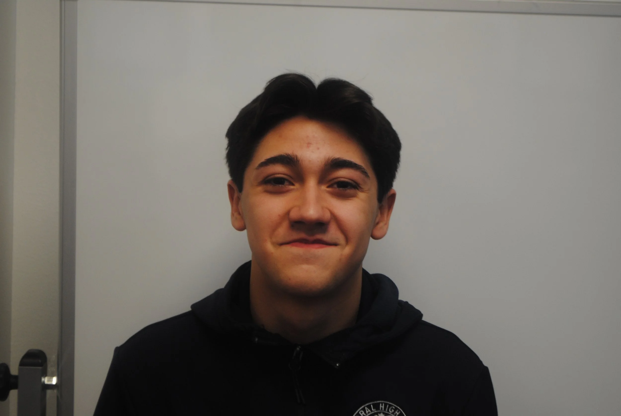 A young man with dark hair and light skin, smiling slightly, wearing a black hoodie, standing against a light-colored wall.