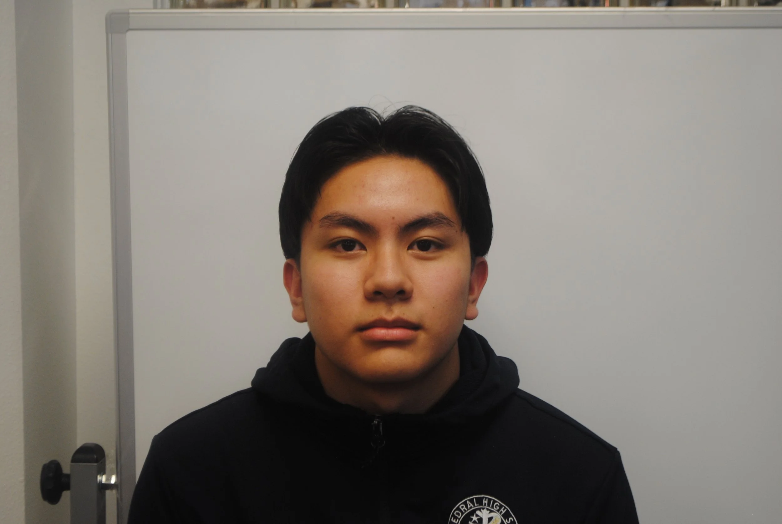 Young man with dark hair, wearing a black hoodie with a logo, sitting against a white background.