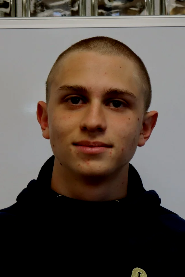 A young man with a short buzz cut hairstyle and wearing a black hoodie, standing in front of a gray wall with a whiteboard behind him.