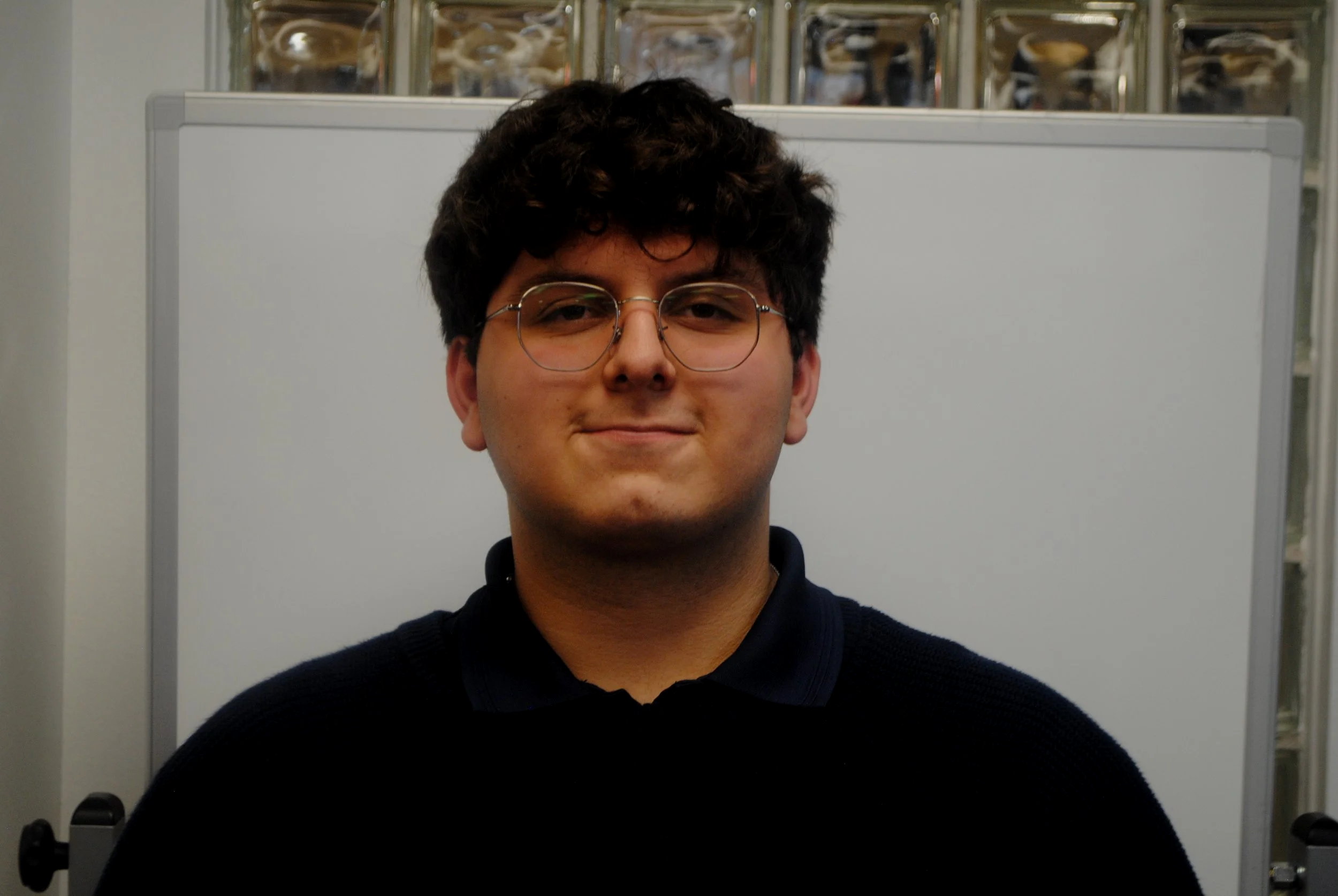 A young man with dark, curly hair and round glasses, wearing a dark sweater, standing in front of a whiteboard and a glass display case.