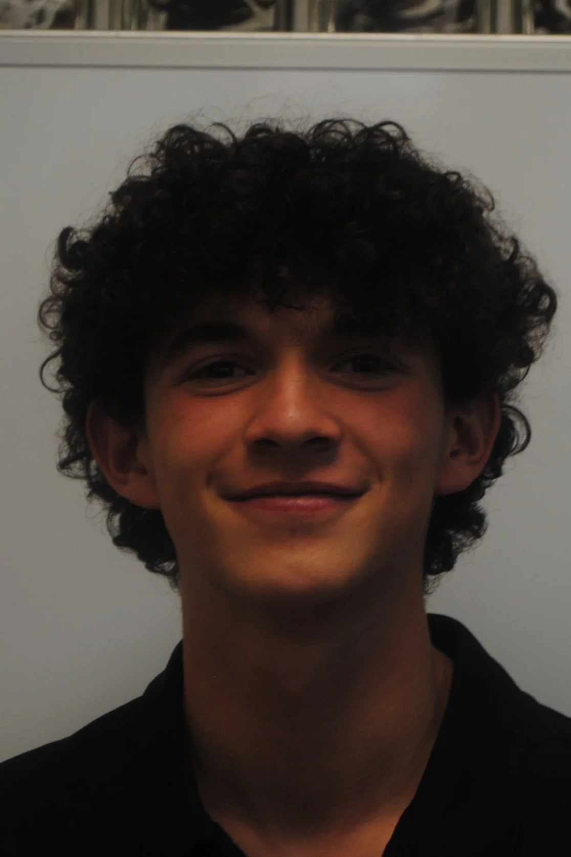 A young man with curly dark hair and a slight smile, wearing a black shirt, standing in front of a plain background.