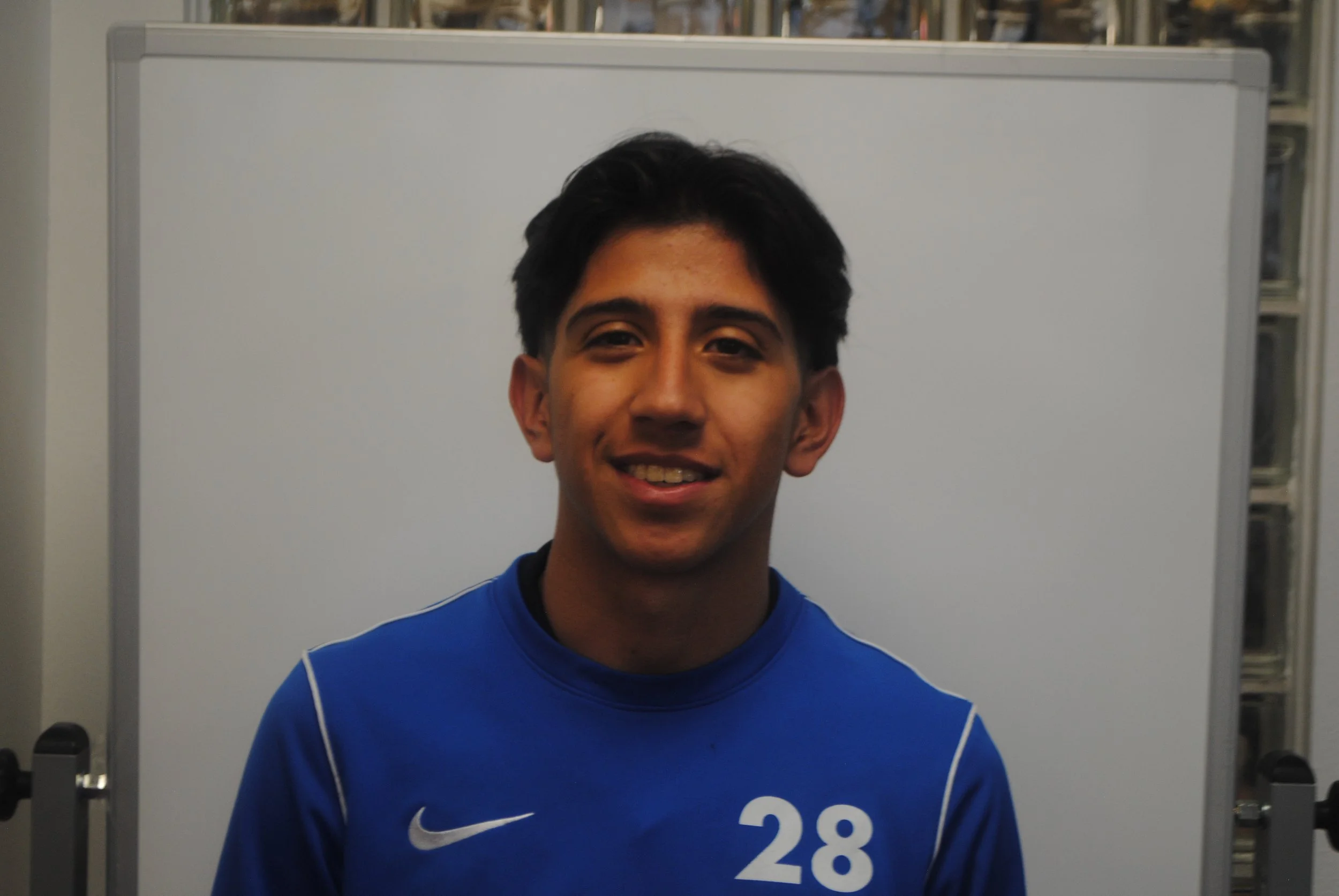 Young man smiling, wearing a blue sports jersey with the number 28, standing in front of a whiteboard.