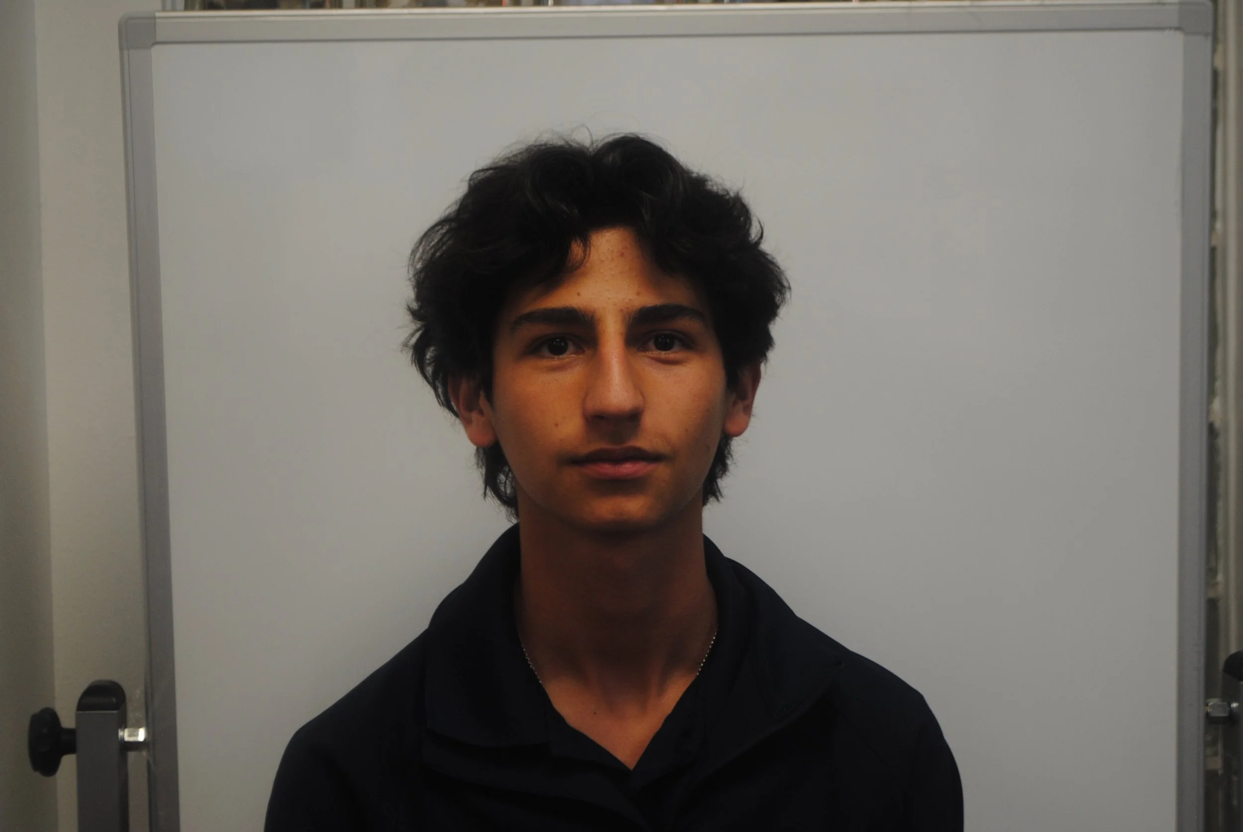 A young man with dark curly hair and light brown skin posing in front of a plain whiteboard.