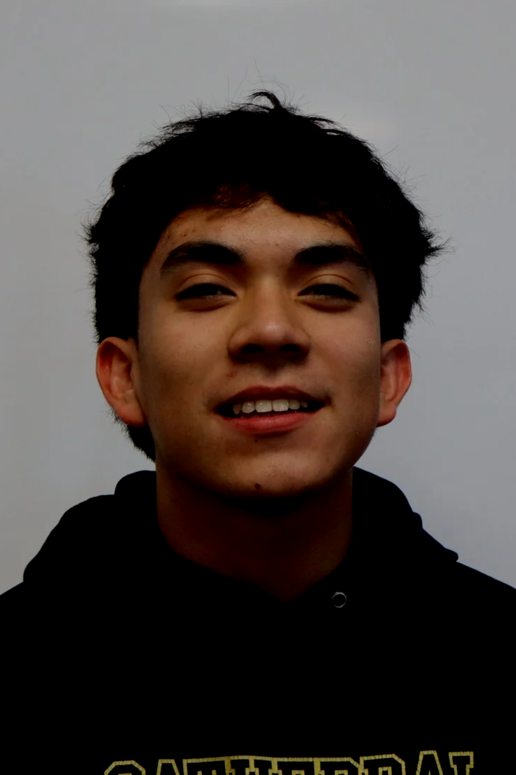 A young man with dark curly hair smiling at the camera, wearing a black hoodie with yellow text, against a plain white background.