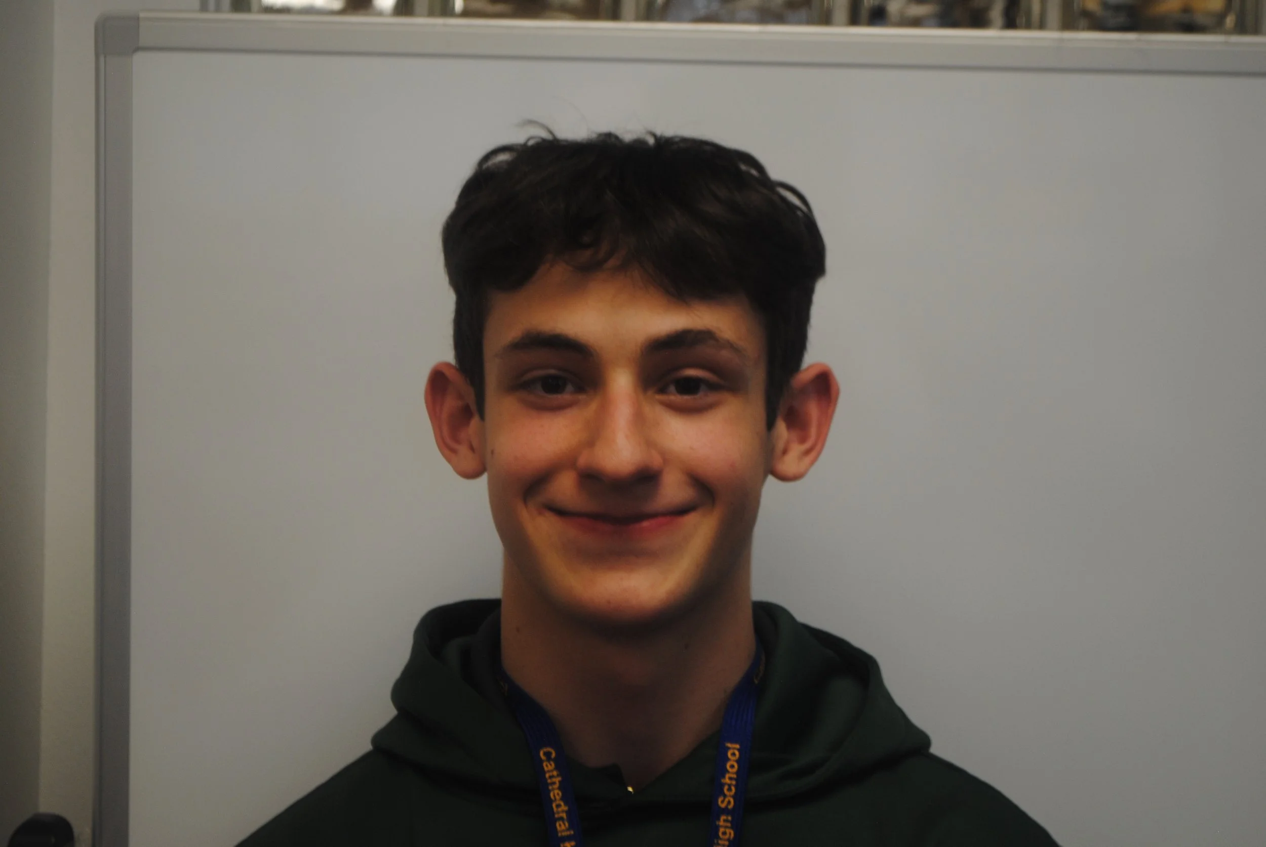 A young man with dark hair, smiling, wearing a black hoodie and a blue lanyard, standing in front of a whiteboard.