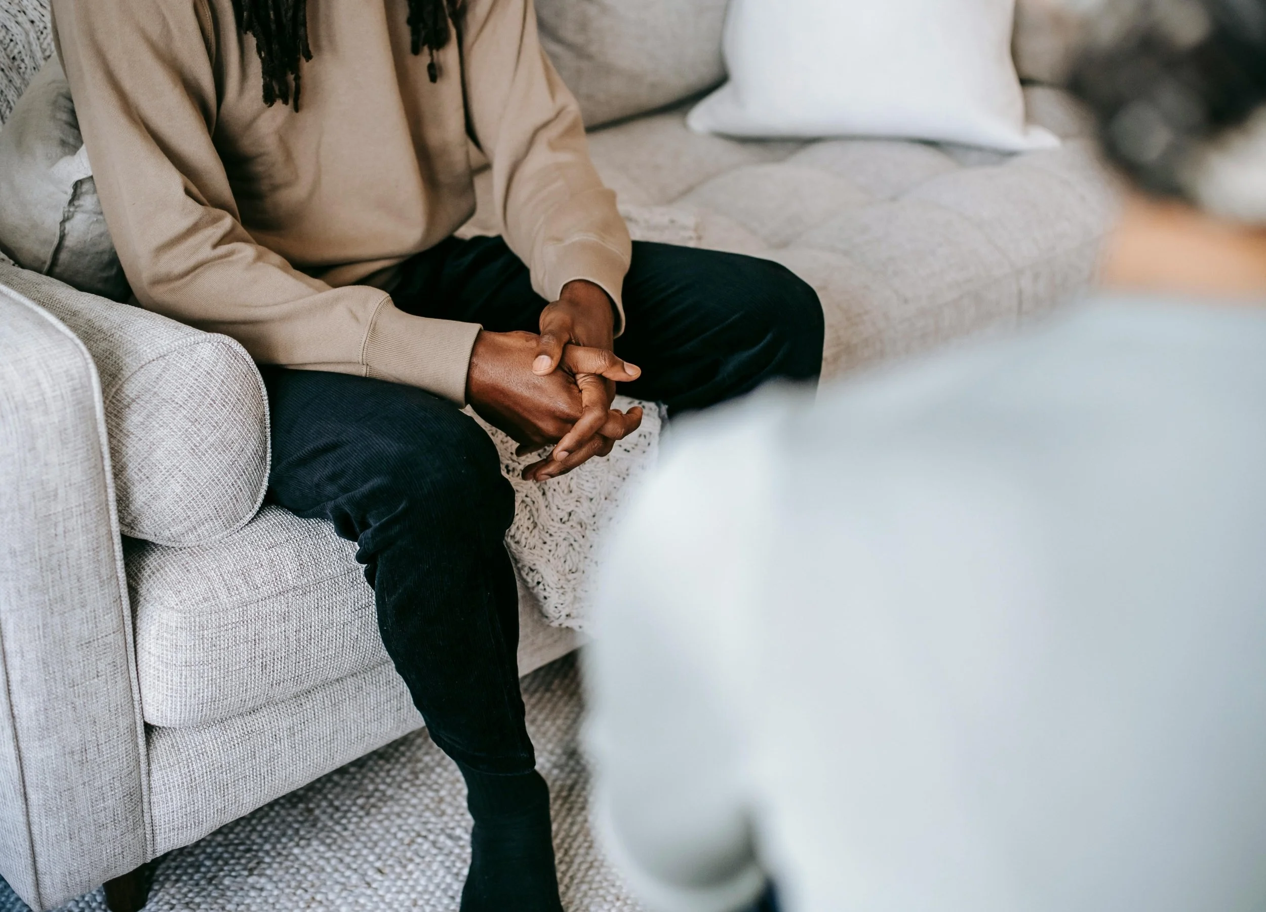 Person sitting on a light gray couch with their hands clasped, engaged in conversation during a therapy session.