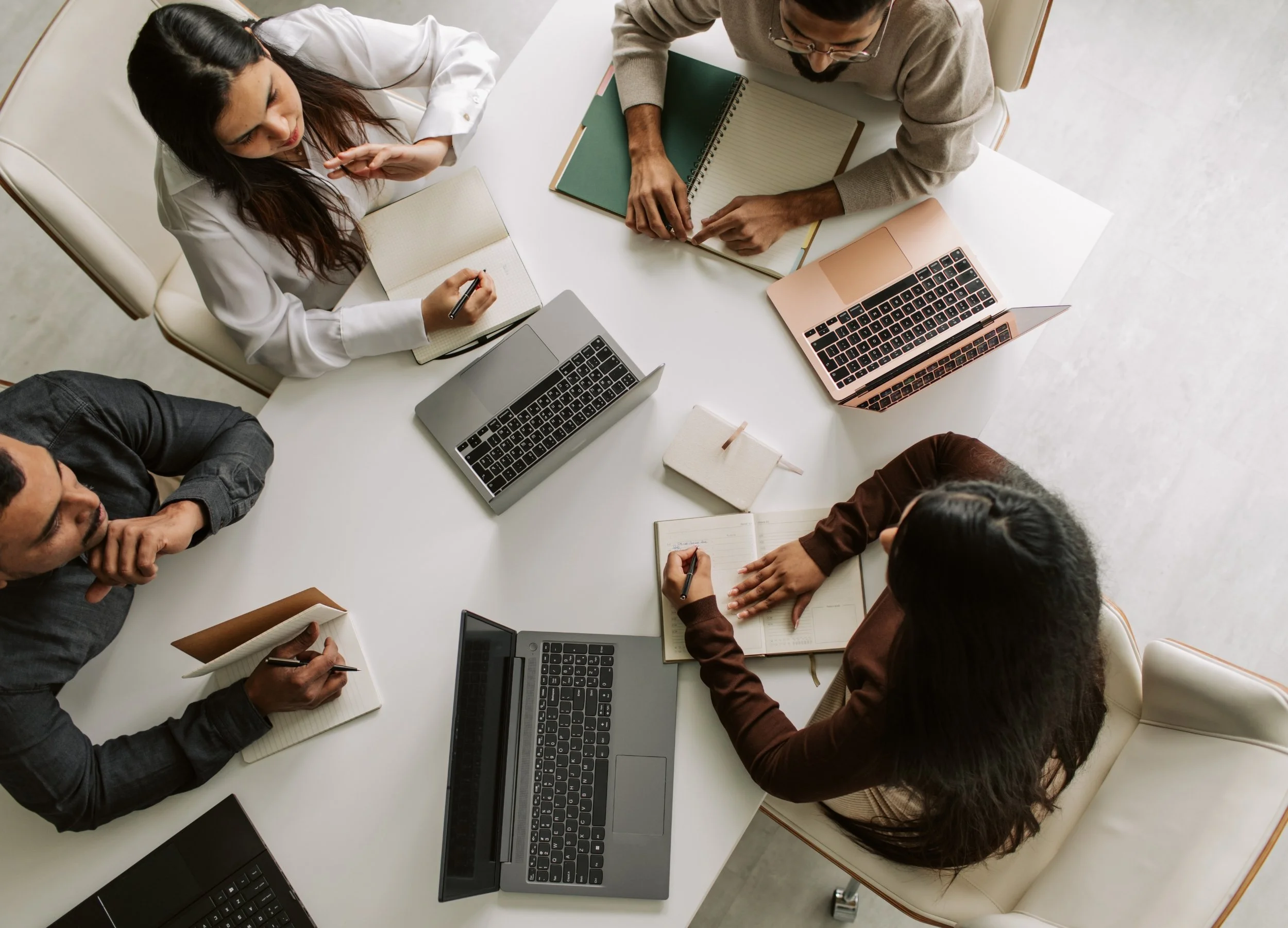 A group of five people sitting around a white round table, working on laptops and taking notes with notebooks and pens, engaged in a meeting or collaborative discussion.