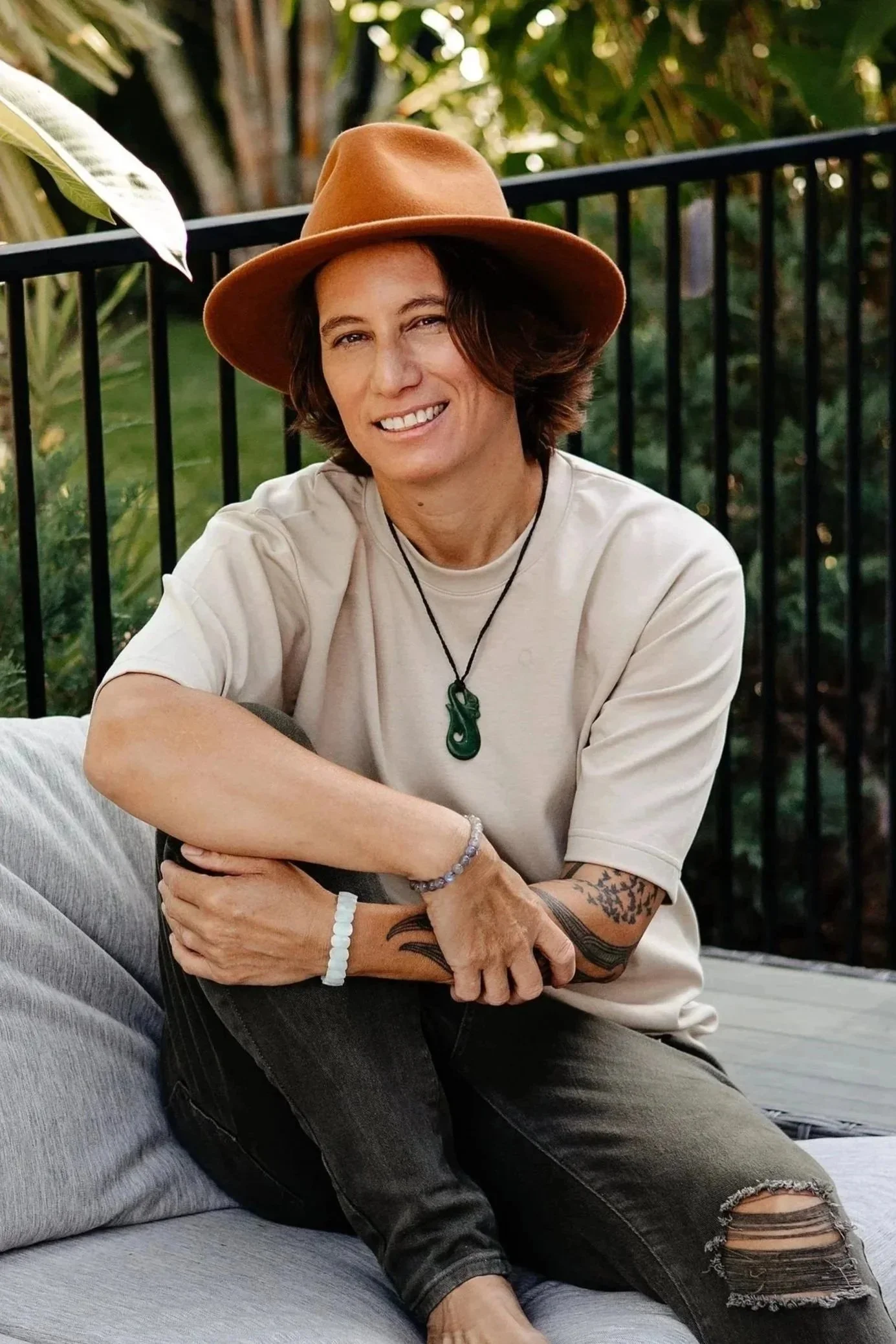 A person with shoulder-length brown hair wearing a tan wide-brimmed hat, a beige T-shirt, and black ripped jeans sitting outdoors against a black metal fence and lush green foliage, smiling at the camera.