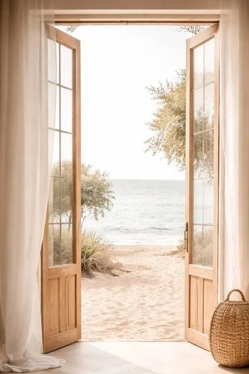 Open wooden door leading to a sandy beach with trees and the ocean in the distance.