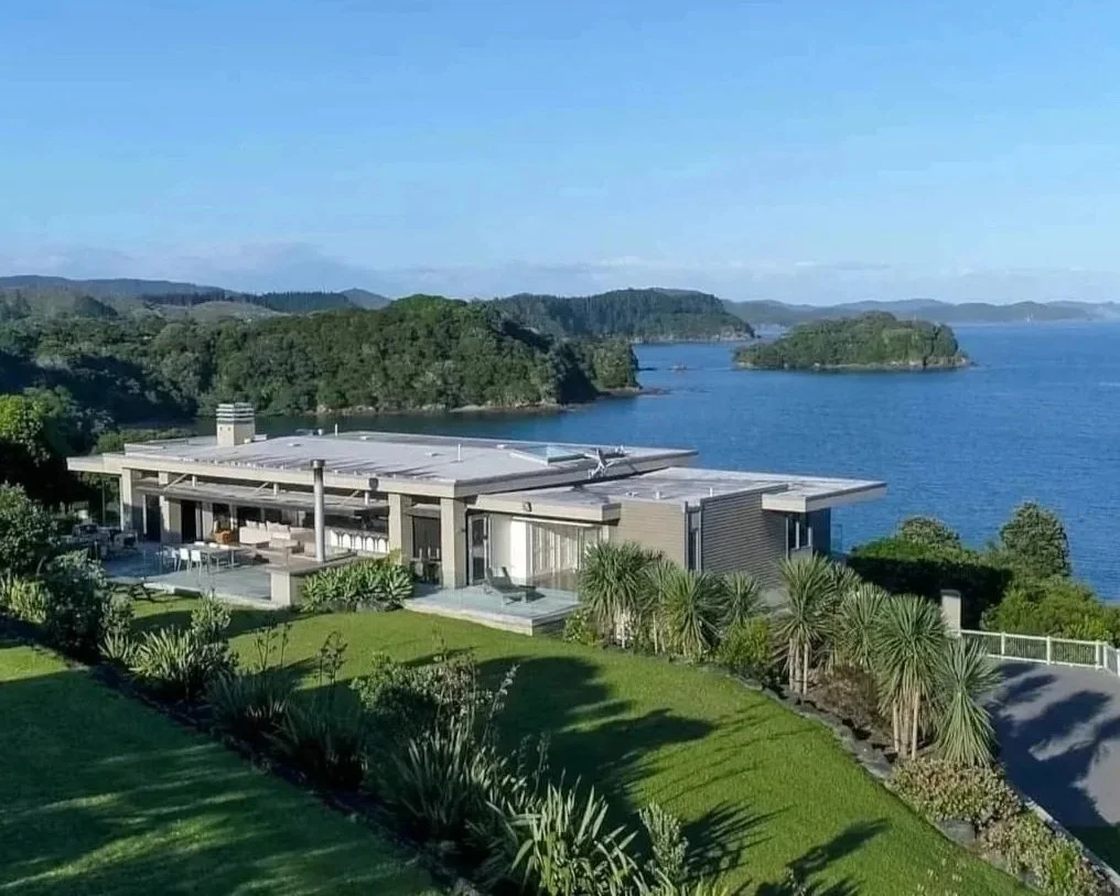 Modern house with flat roof and large windows on a hill overlooking a body of water with small islands and a forested shoreline in the background.