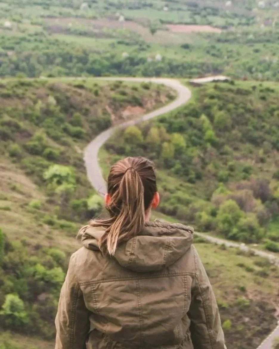 Woman with brown hair tied in a ponytail wearing a brown jacket, standing and looking at a winding road through green hilly landscape.