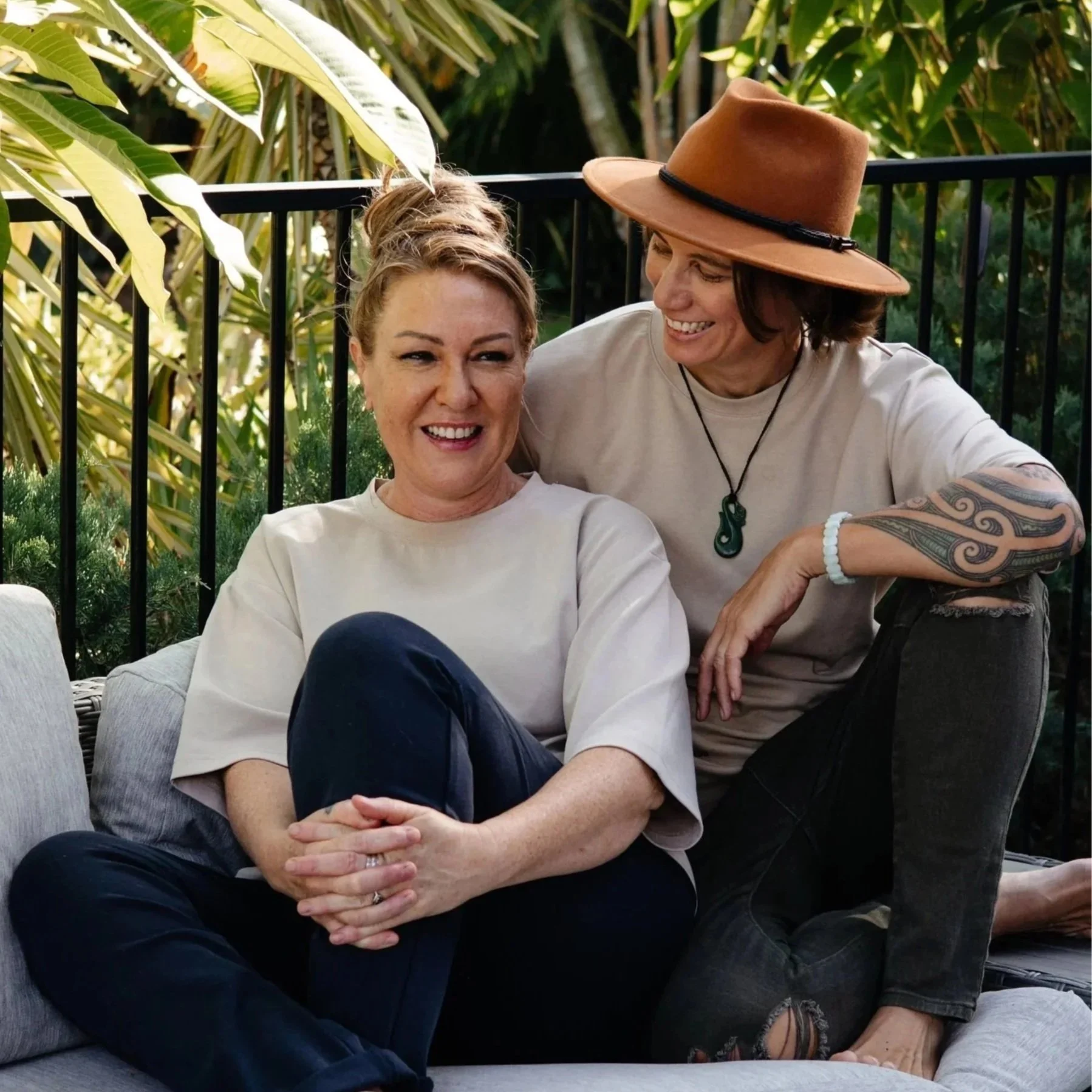 Two smiling women sitting closely together outdoors, one with a brown wide-brimmed hat and tattooed arm, the other with blonde hair styled in an updo, surrounded by green plants and a black metal fence.