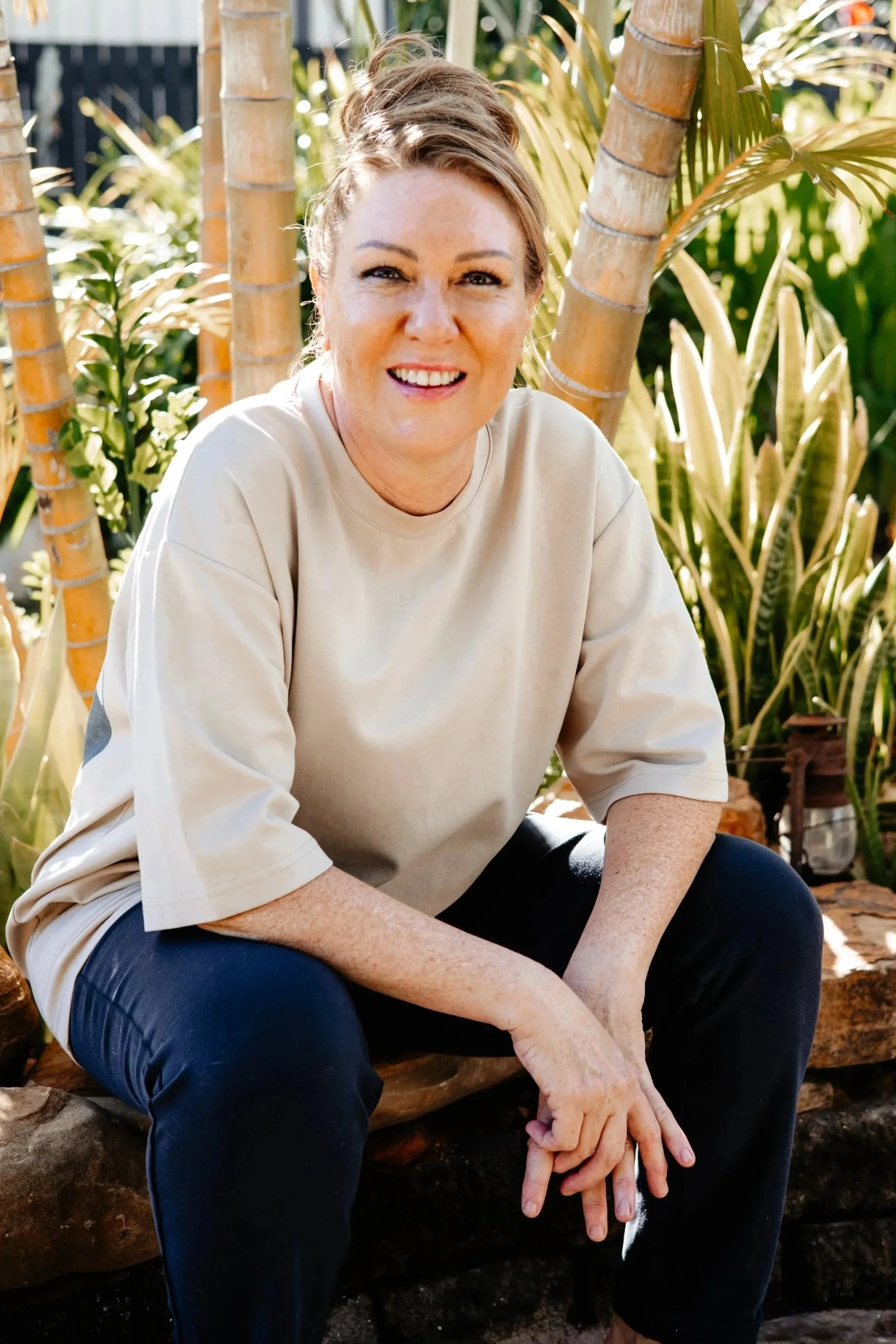 Woman with red hair, freckles, and a beige shirt sitting outdoors among green plants and palm trees, smiling at the camera.