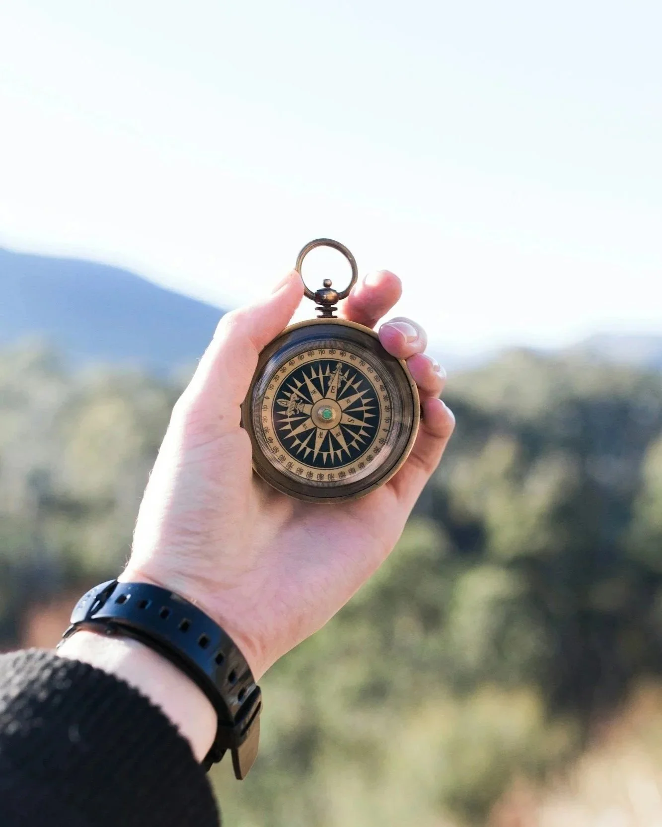 A person holding a vintage compass outdoors with a background of blurred natural scenery and mountains.
