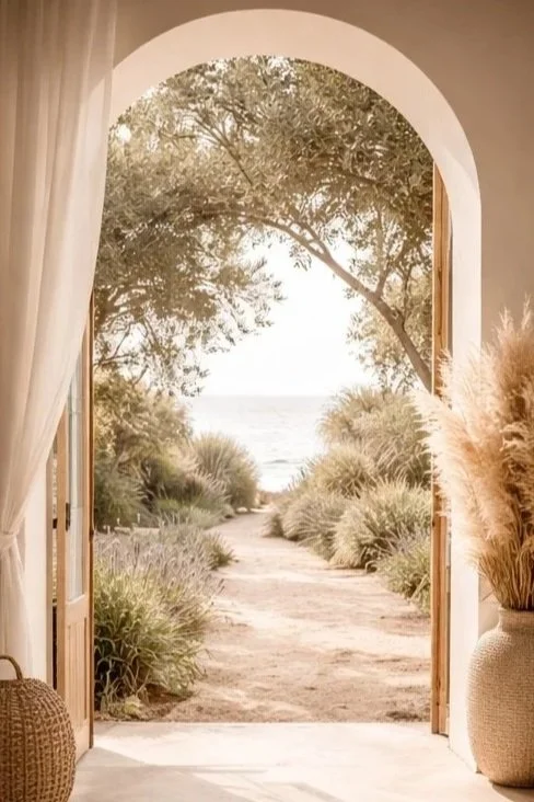 Open door opens to a beach path with trees and bushes leading to the ocean at sunset or sunrise.