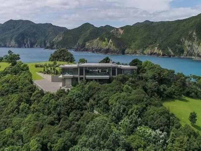 Modern house on a hillside overlooking a body of water, surrounded by lush green trees and mountains in the background.