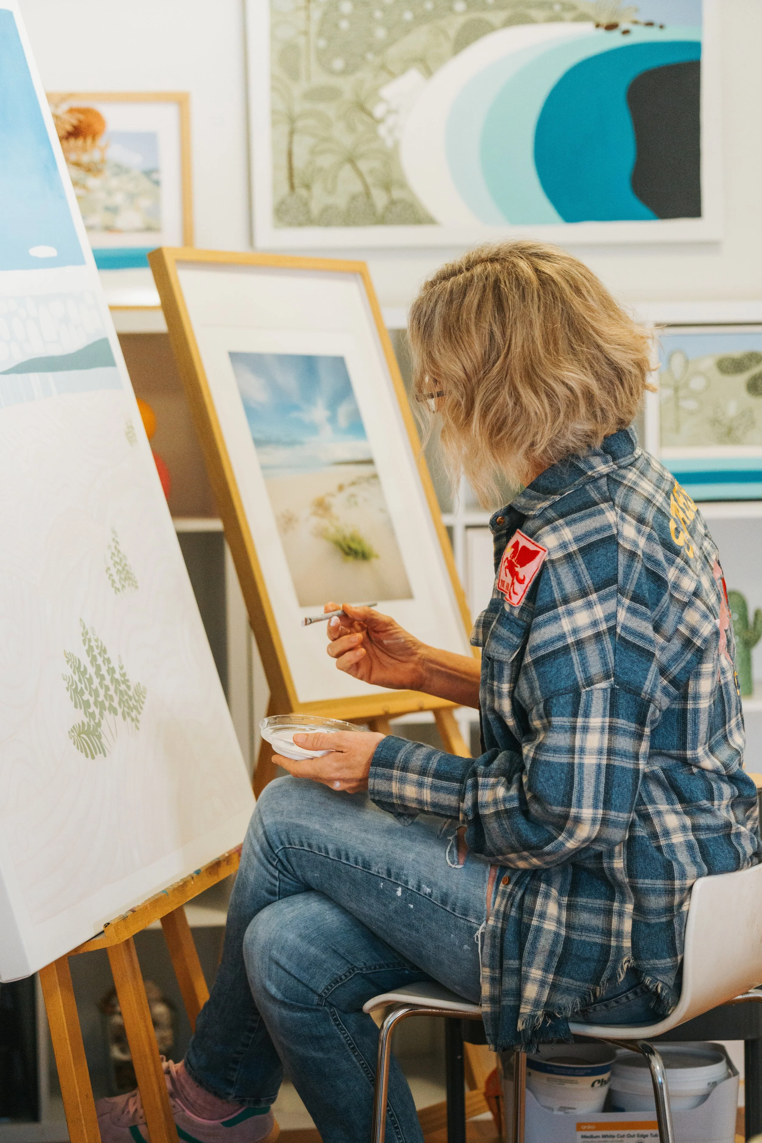 A woman with curly hair and glasses, dressed in a plaid shirt and jeans, is sitting on a chair, painting on a large canvas with trees. She is holding a cup and a small palette. Several framed landscape paintings hang on the wall behind her.