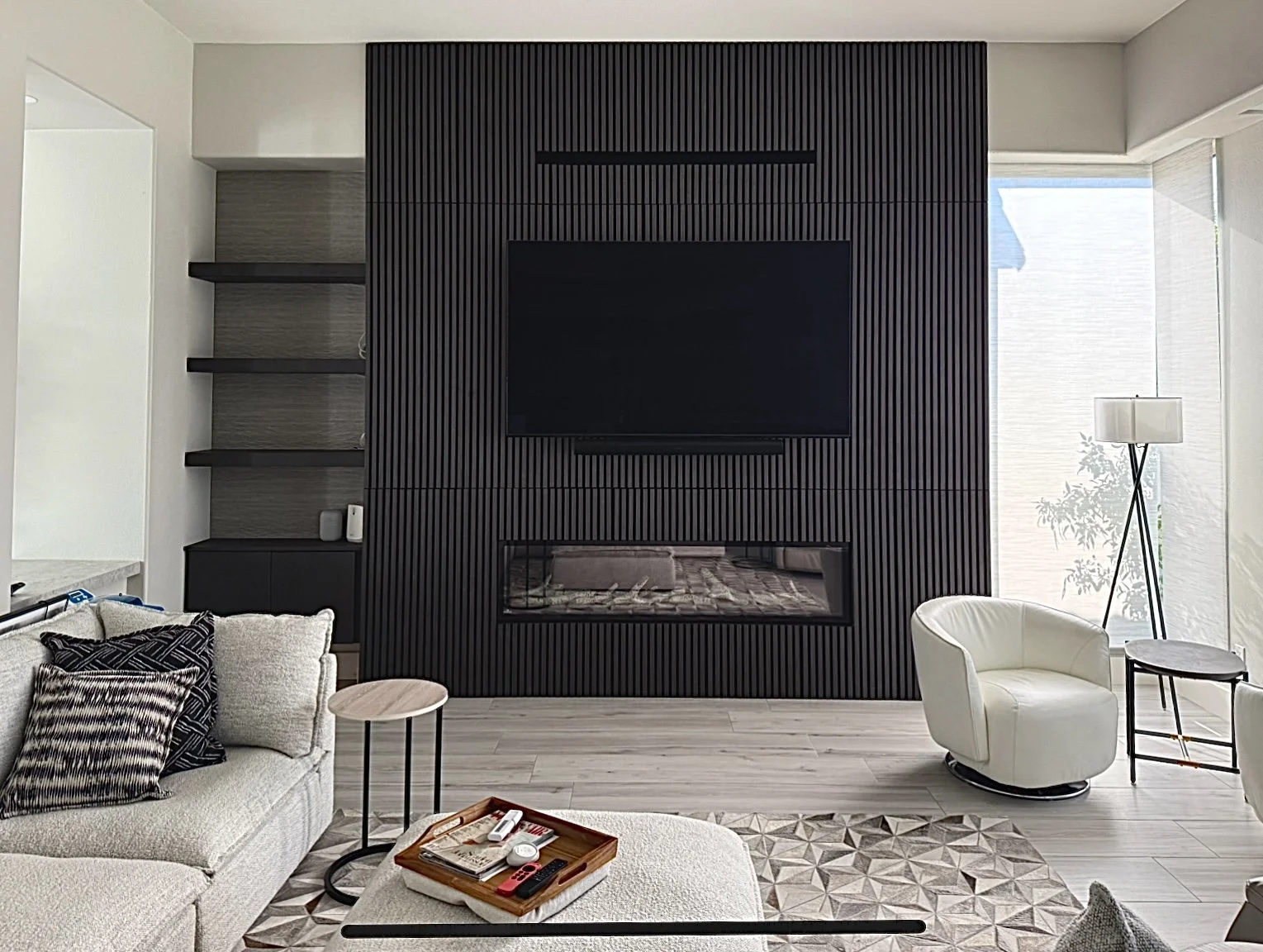 Modern living room with beige sofa, black accent pillows, small side tables, white armchair, fireplace, flat-screen TV on a black textured wall, bookshelf, large window with natural light, and a floor lamp.