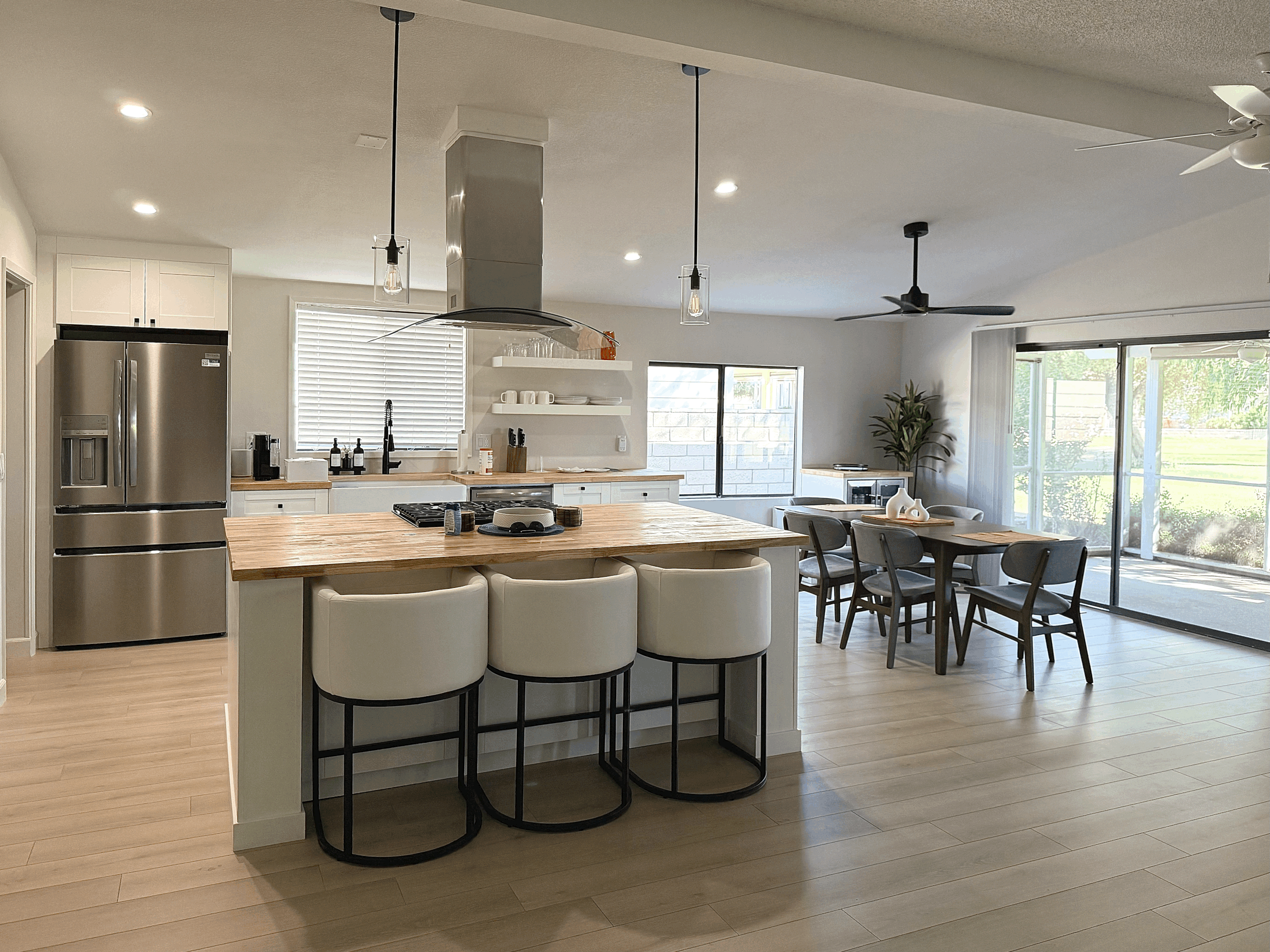 Open-concept kitchen and dining area with white cabinetry, stainless steel refrigerator, wooden island with seating, white barstools, dining table with chairs, large sliding glass door, and hardwood floors.