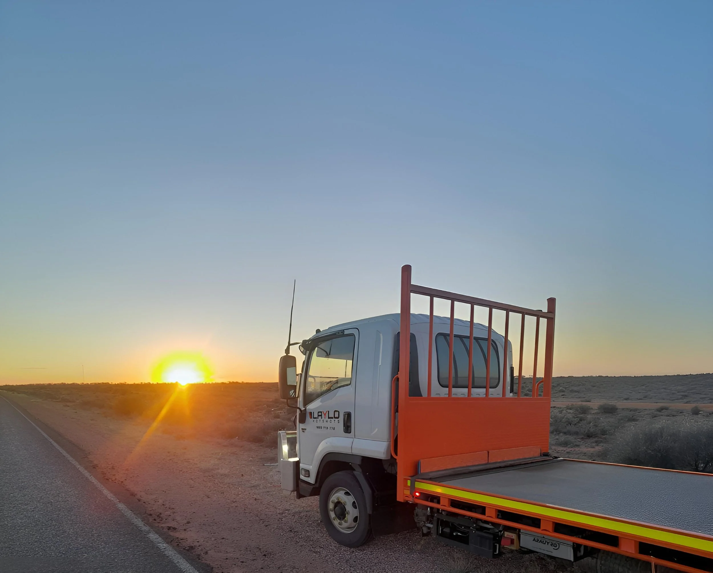 A white utility truck with orange and yellow safety markings on the side, parked beside a road at sunset with a clear sky and distant mountains in the background.