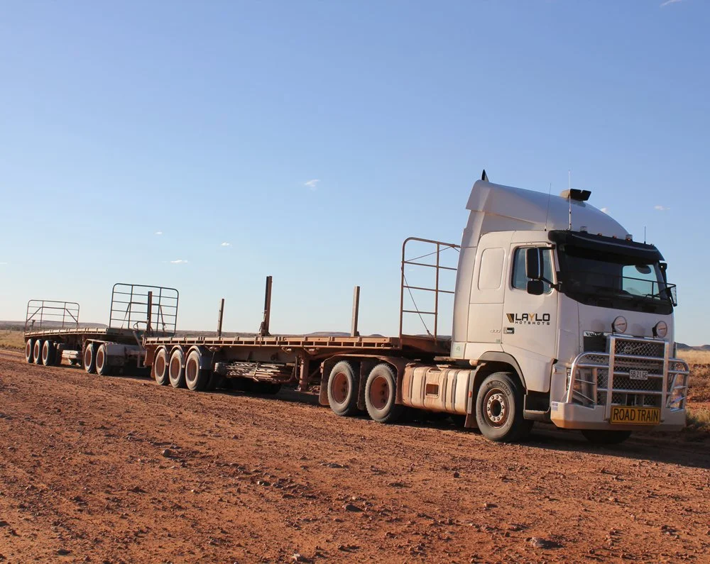 A large white semi-truck with a flatbed trailer parked on a dirt road in a rural desert area under a clear blue sky.