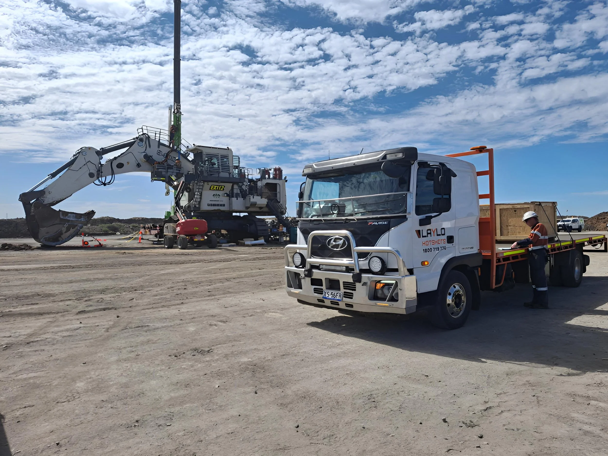 Construction site with a large excavator lifting a green pole, and a white Hyundai truck with the branding 'LAYLO' parked nearby under a blue sky with scattered clouds, with a worker wearing a white helmet and orange vest nearby.
