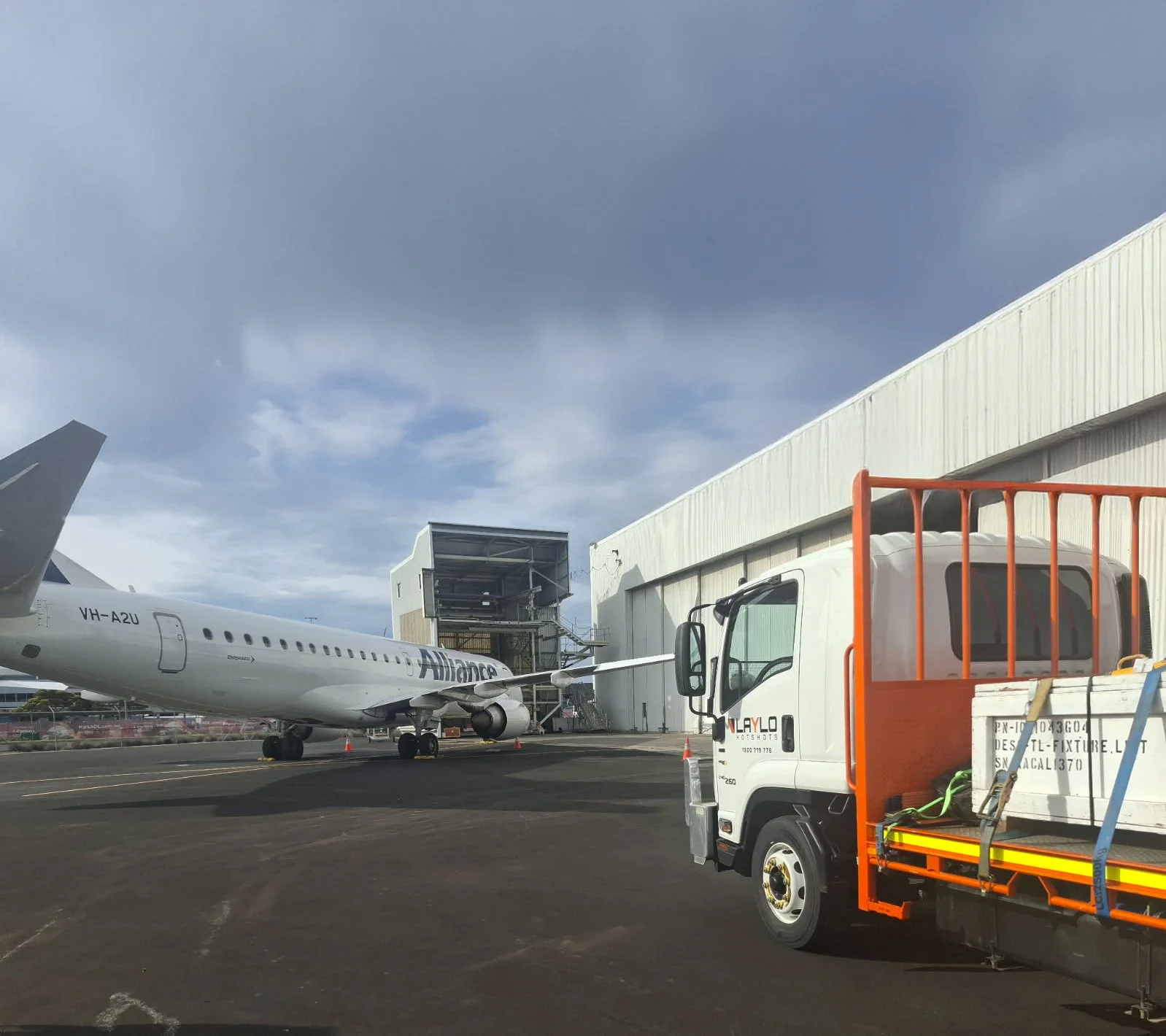 An airplane on the tarmac with a cargo truck nearby and a warehouse in the background. The sky is mostly cloudy.