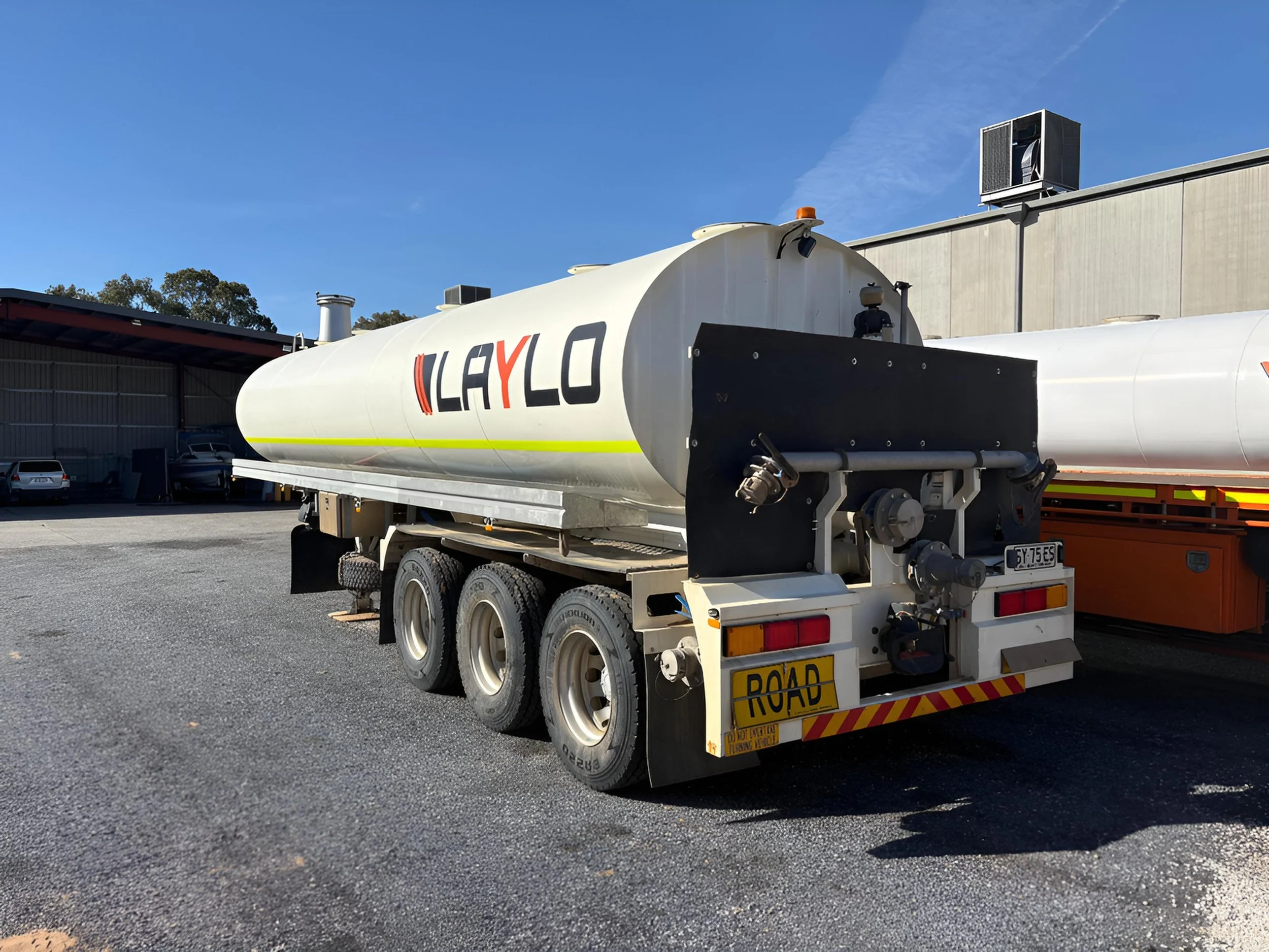 Large white tanker truck with yellow and red accents parked outdoors near a building, with trees and a clear blue sky in the background.