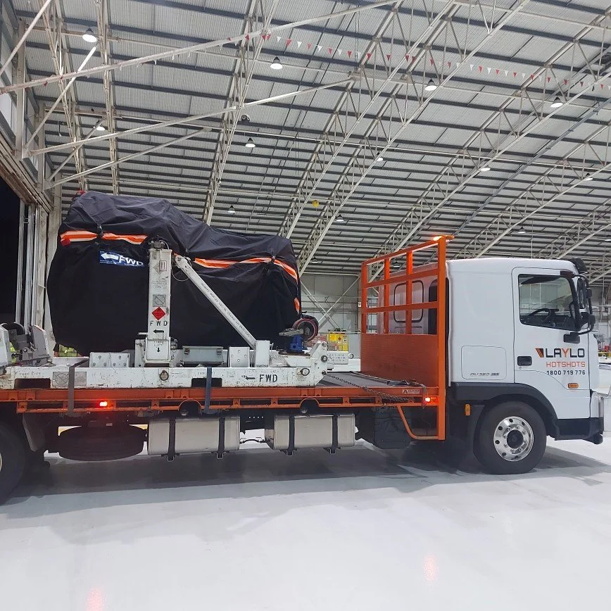 A white flatbed truck with orange side rails inside a large warehouse. The truck is carrying a loaded cargo secured with black cover and orange straps.