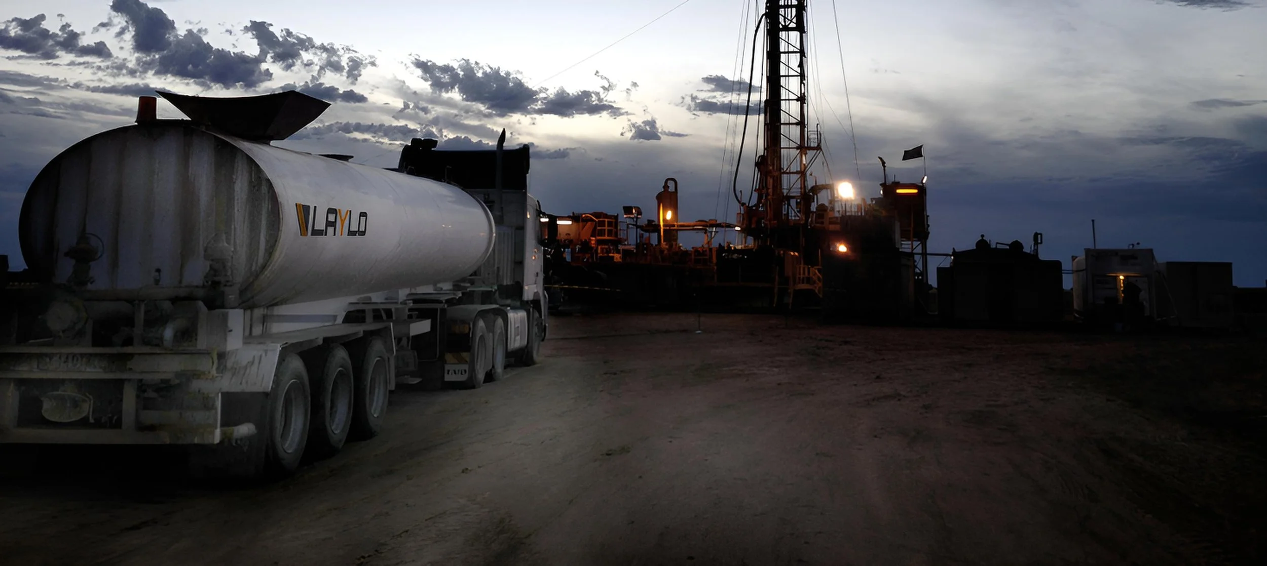 An industrial scene at dusk or dawn featuring a large white tanker truck labeled 'VLAYD' on the left, and heavy machinery and equipment with lights on, including a tall crane structure, against a cloudy sky.