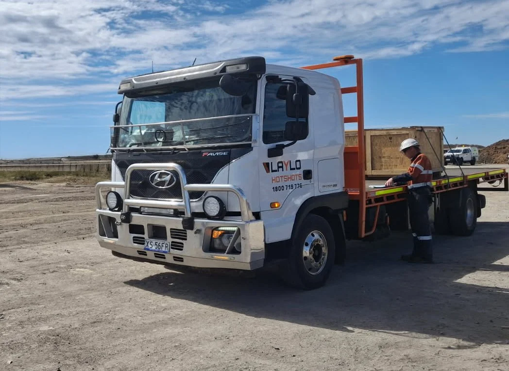 Construction worker in safety gear standing next to a Hyundai flatbed truck on a dirt lot under a partly cloudy sky.