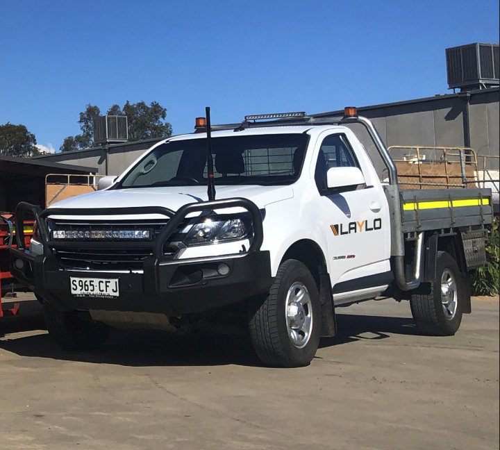 A white utility truck with a black front bumper and grille guard, marked 'LAYLO' on the door, parked outside on a concrete surface under a clear blue sky.
