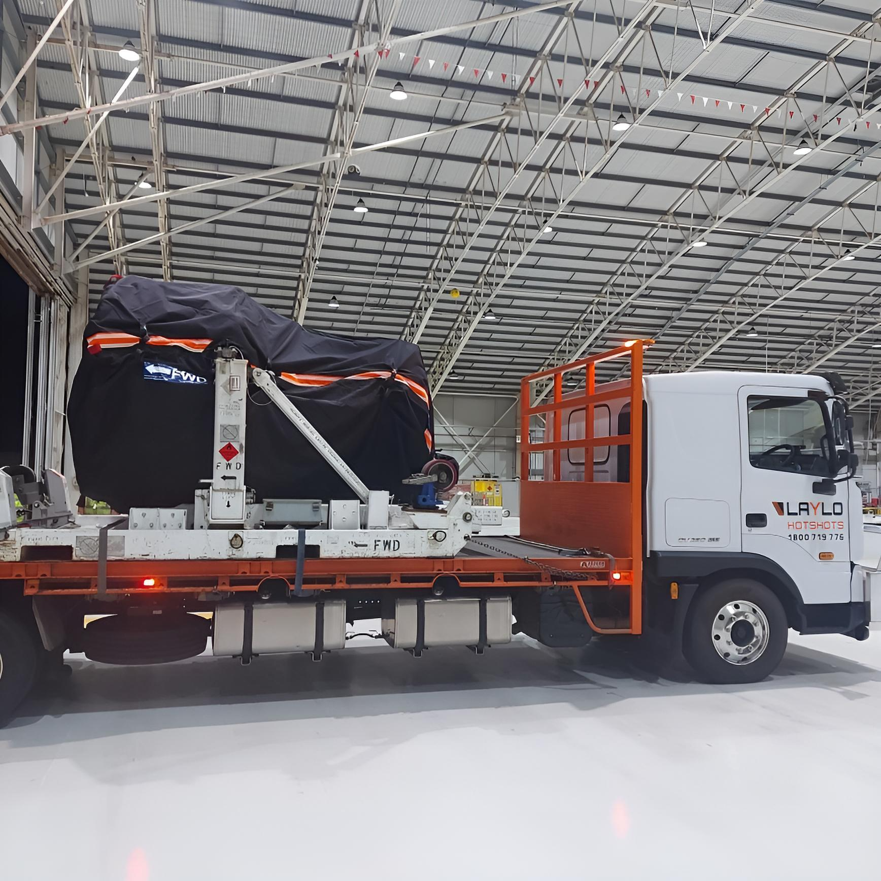 A white truck inside a warehouse with a high curved metal roof. The truck is carrying a large covered load on a flatbed trailer. The load is wrapped in black plastic with orange accents. The warehouse has bright lighting with several ceiling lights, and small red and white flags hang from the roof structure.