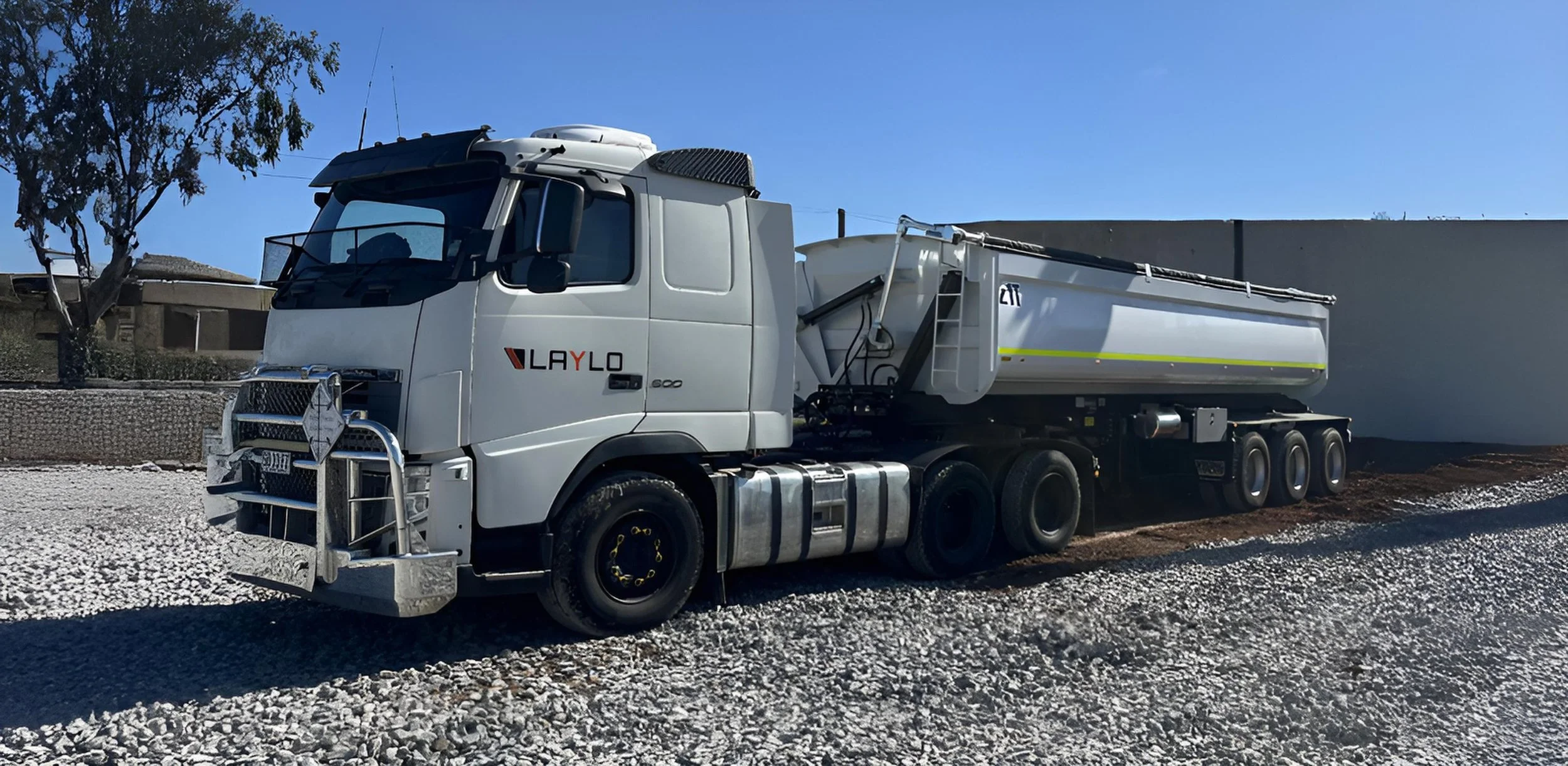 A white Laylo fuel truck parked on gravel under a clear blue sky with a building and trees in the background.