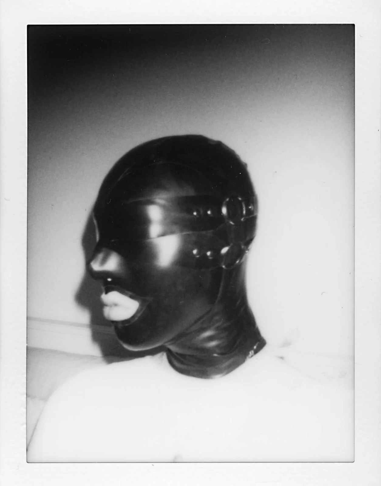 Black and white photo of a mannequin head wearing a latex mask that covers the entire face, with a shiny and smooth surface, featuring large lips and detailed eye and nose holes.