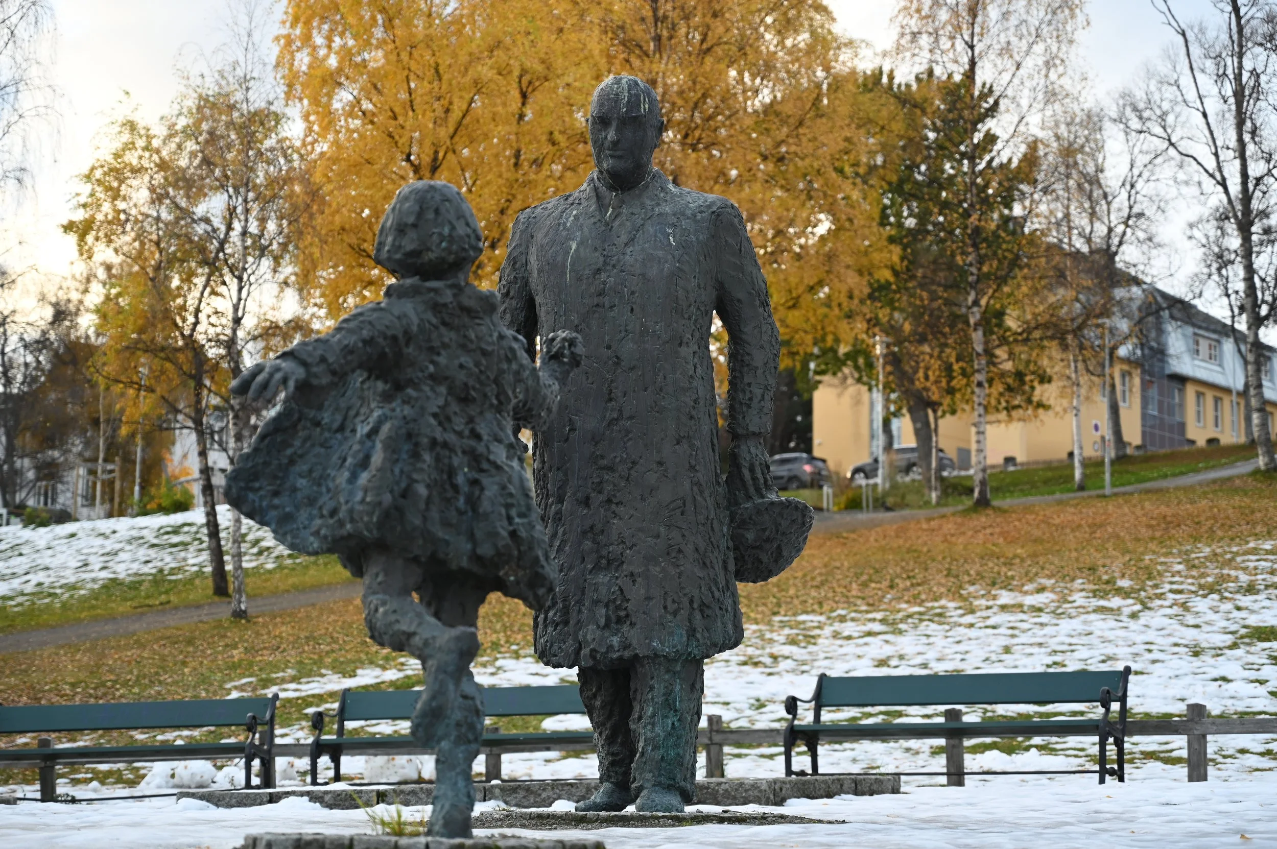 Bronze sculpture of a man in a coat holding a bag, standing and facing a girl in a dress with short hair, in a park with fallen snow, leafless trees, and autumn-colored trees in the background.