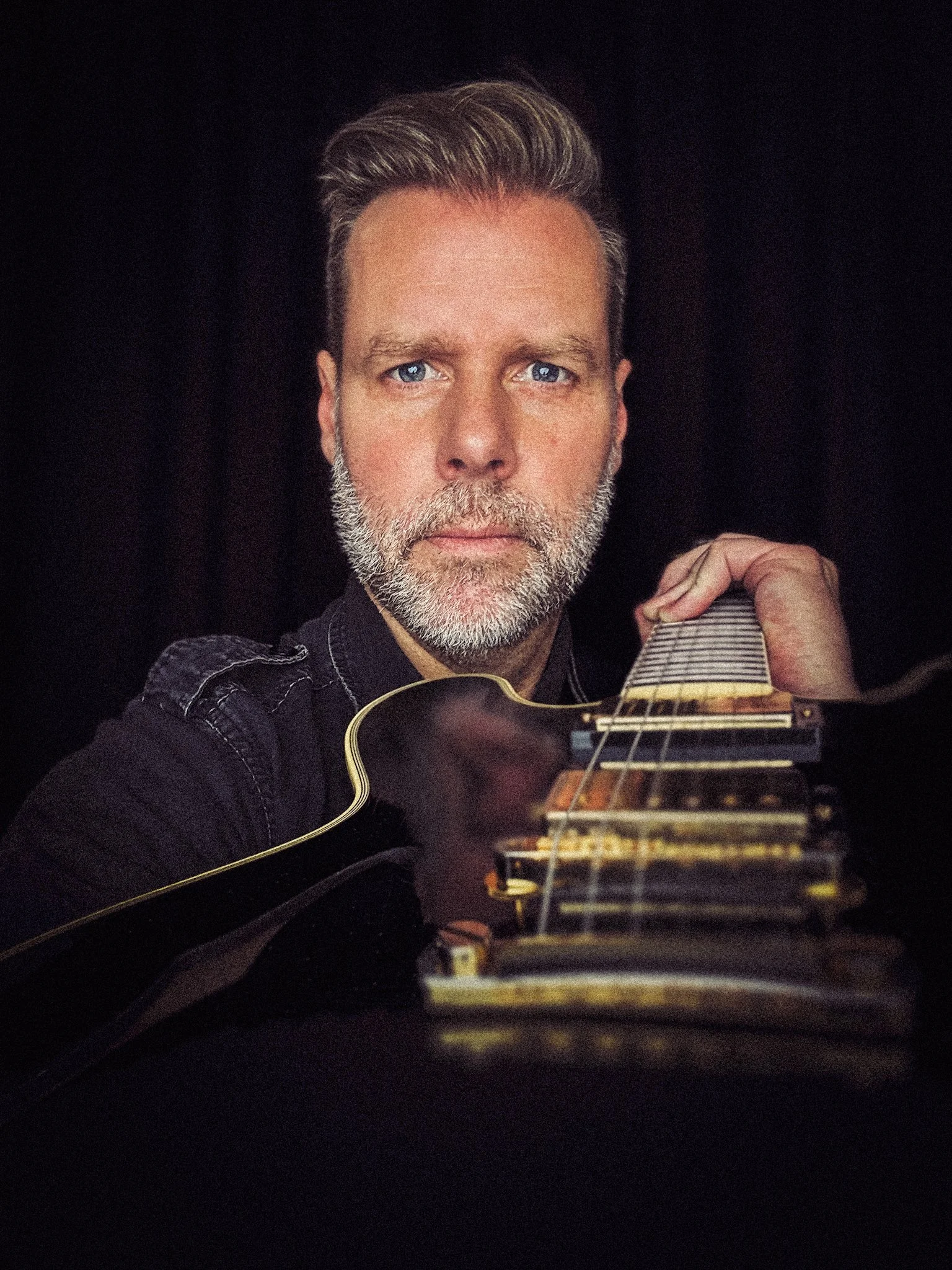 Close-up of a man with gray hair and beard holding an acoustic guitar, looking directly at the camera, against a dark background.