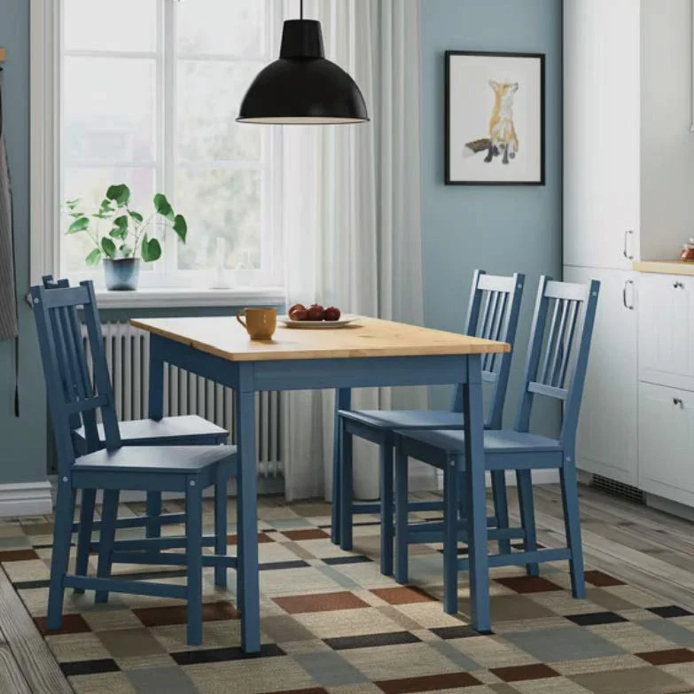 A dining area with a wooden table painted blue, surrounded by three matching blue chairs. On top of the table, there is a small orange mug and a plate with apples. A large black pendant light hangs above the table. In the background, there is a window with white curtains, a green potted plant, a framed picture of an animal on a blue wall, and part of a white kitchen cabinet.