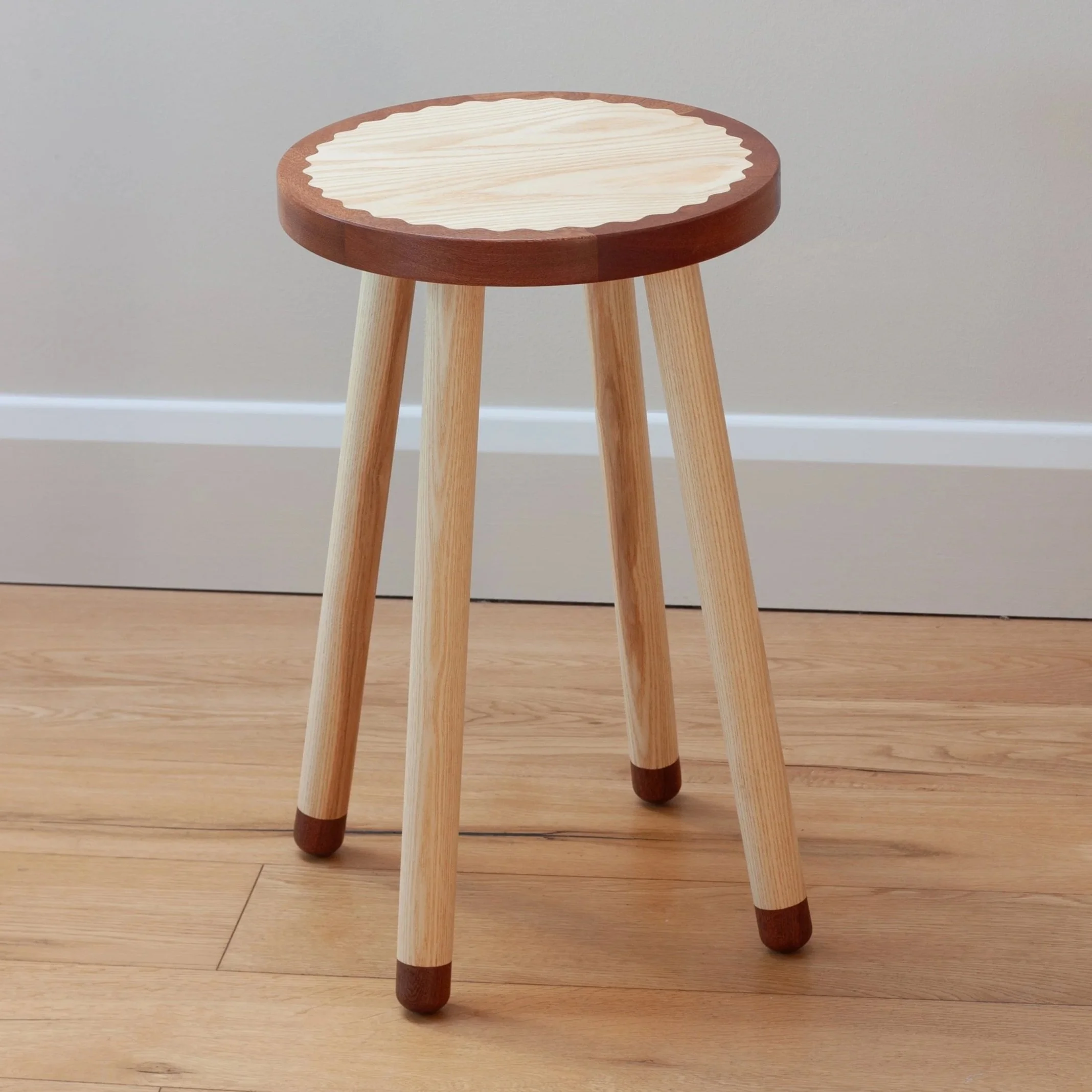 A wooden stool with four legs, with a round seat featuring a two-tone wood pattern and a scalloped edge, stands against a plain wall on a wooden floor.