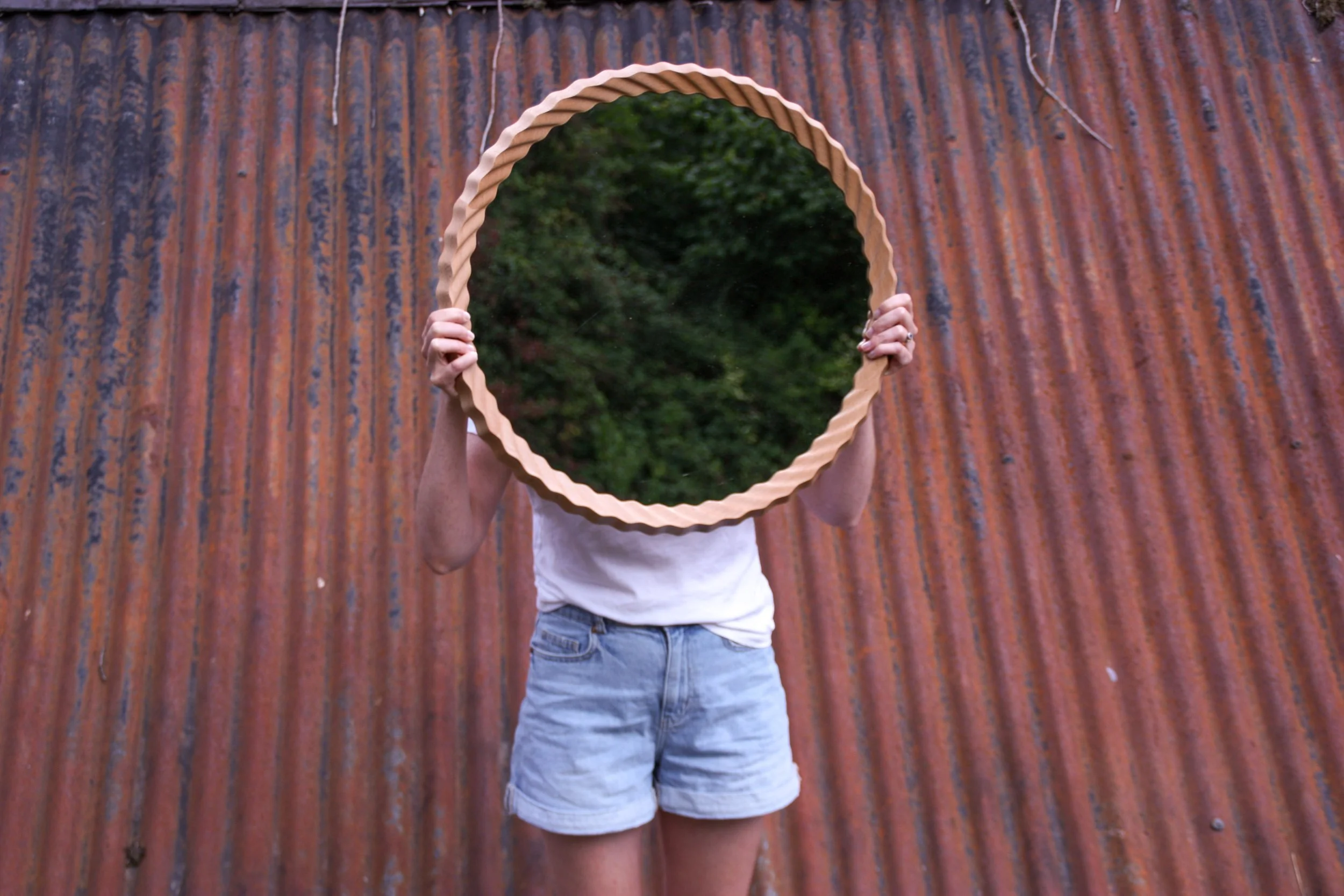 Person standing in front of a rusted metal wall, holding a round mirror with a scalloped edge in front of their face, obscuring their identity. The person's arms are visible, but their face is hidden by the mirror.