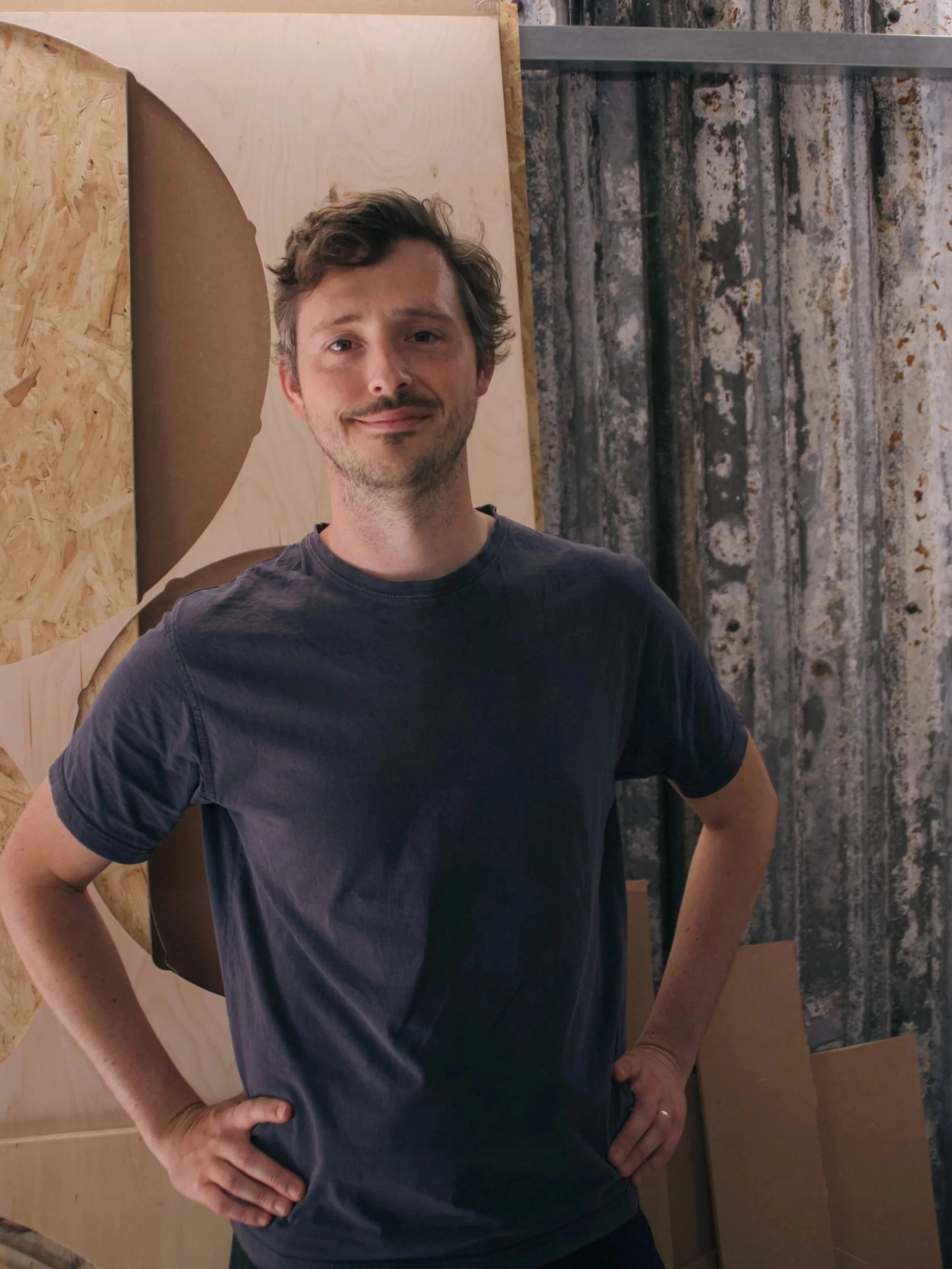 A man with brown, curly hair and a slight beard, wearing a dark t-shirt, standing in front of a plywood and metal textured background with his hands on his hips and a slight smile.
